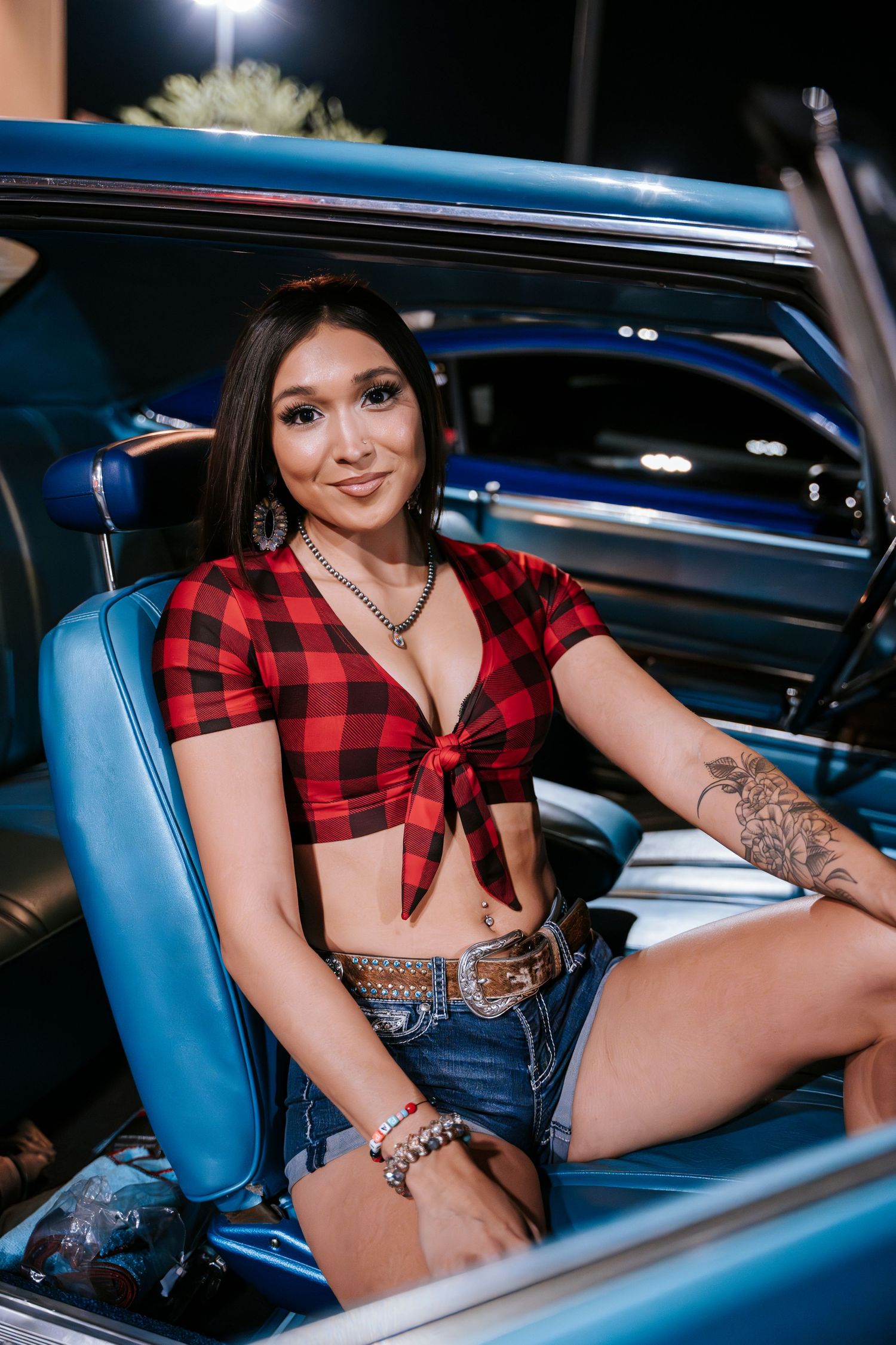 Model in red plaid crop top poses inside classic blue car with vintage interior.