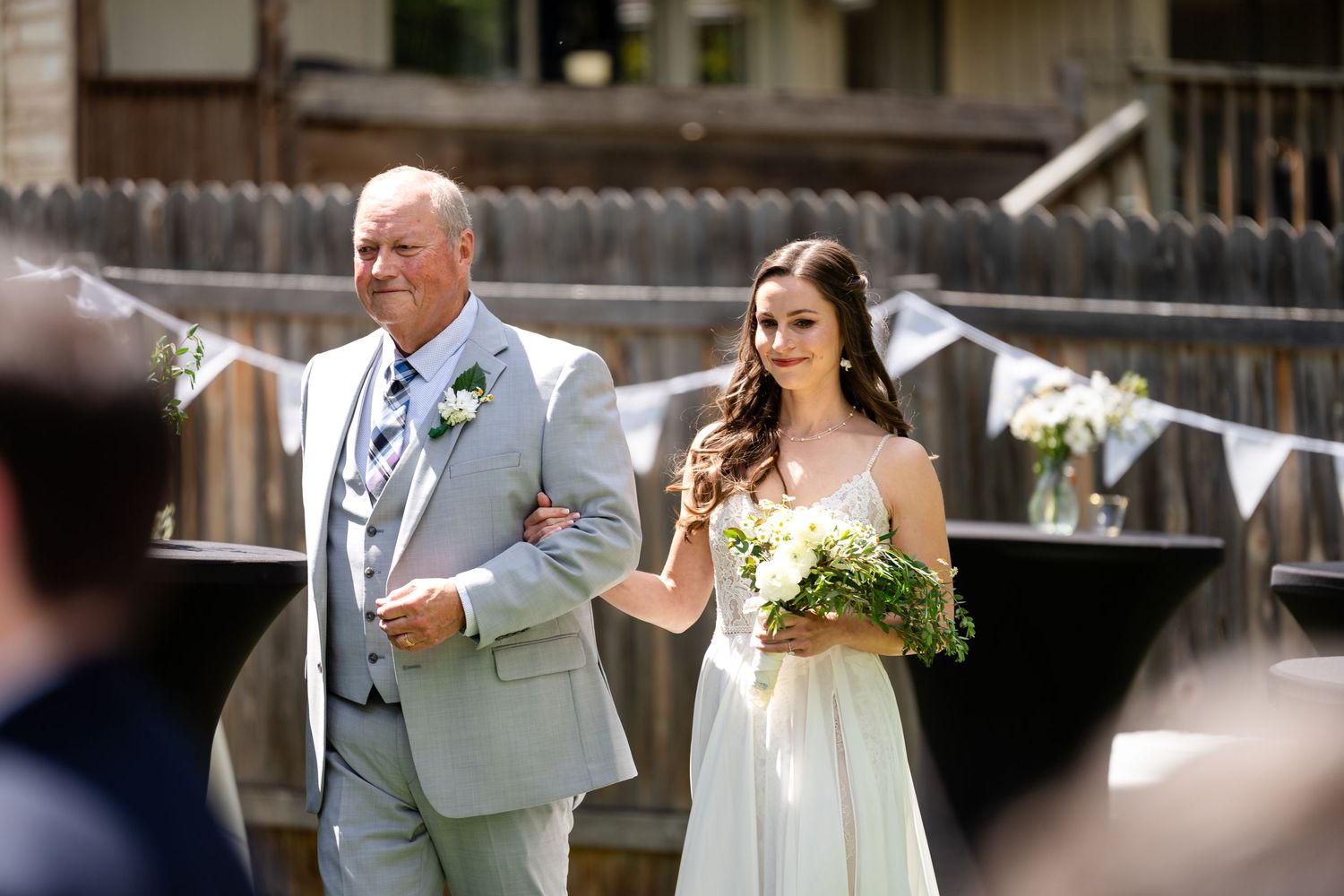 Bride in white gown walks down aisle with escort at outdoor ceremony.