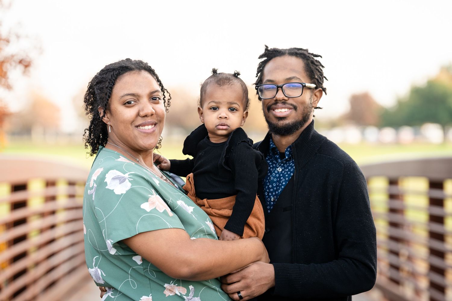 Family of three poses together on wooden bridge during outdoor portrait session.