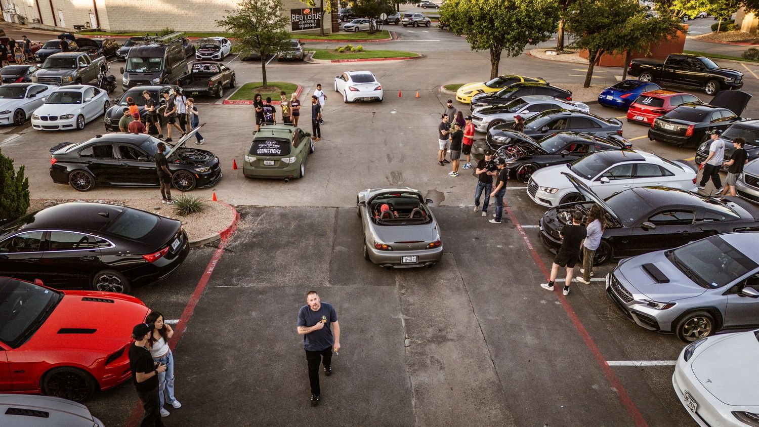 Aerial view of a car meet gathering in a parking lot with various vehicles and attendees.