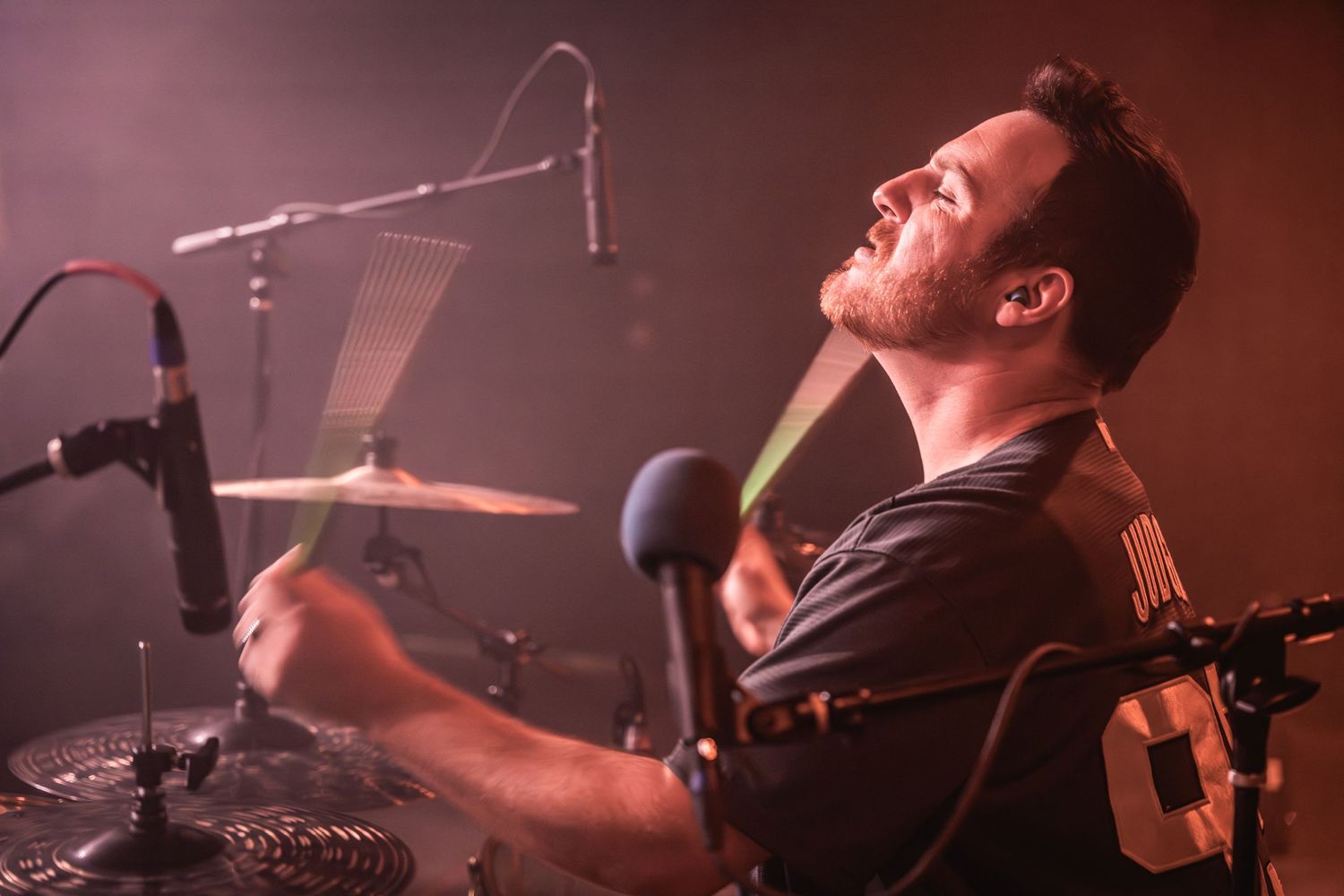A drummer performs passionately behind a drum kit under warm red stage lighting.