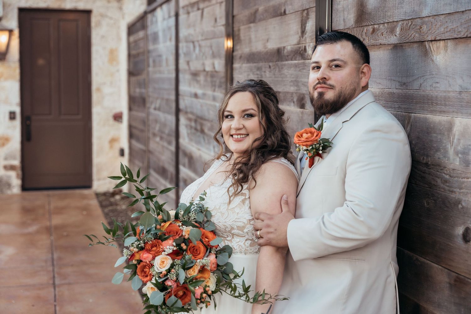 A wedding couple poses against a rustic wooden wall with a coral floral bouquet.