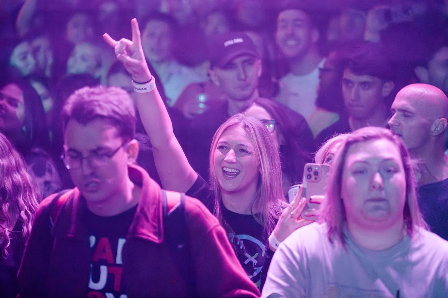 Concert crowd cheering with purple lighting and raised hands at an energetic live music event.