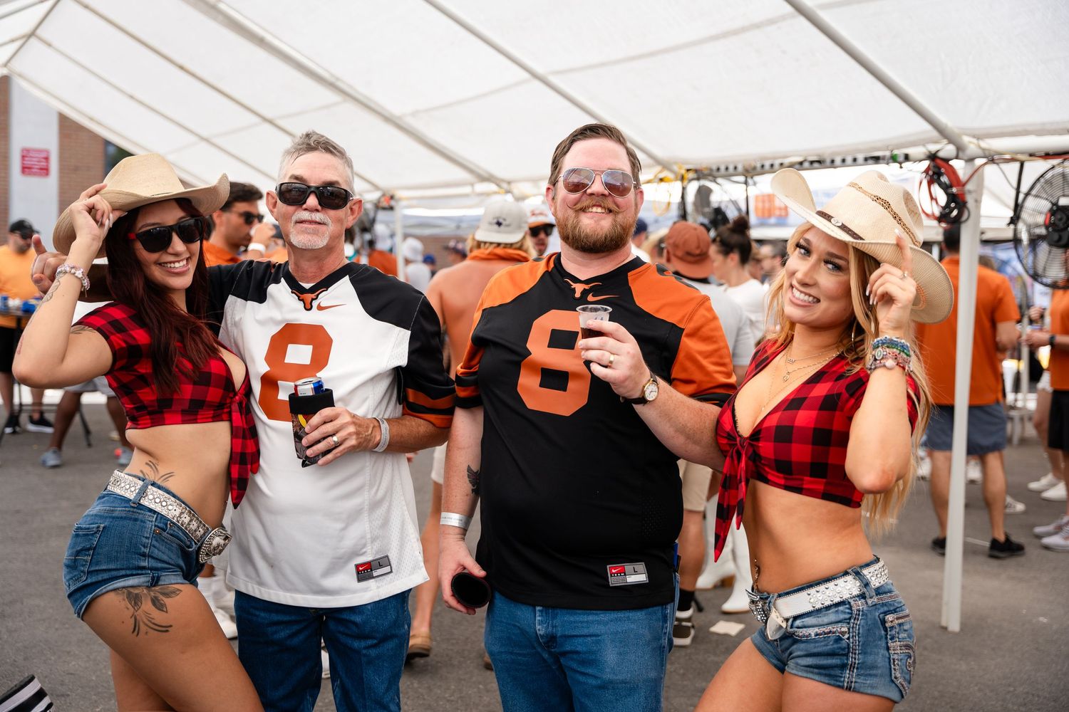 Festival attendees in orange sports jerseys and casual wear pose with drinks under white tent.