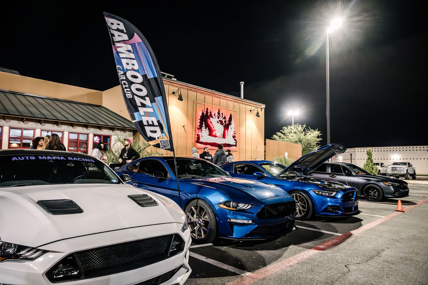 Performance cars lined up at night outside a building with blue flag banner and bright lighting.