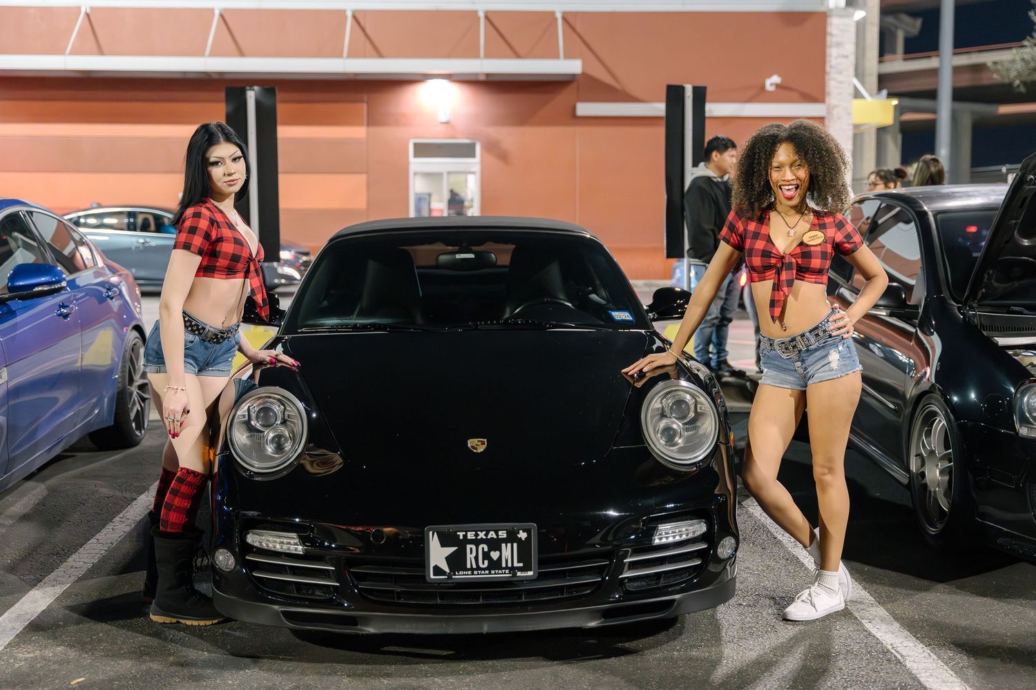Two models pose with black Porsche at nighttime car meet.
