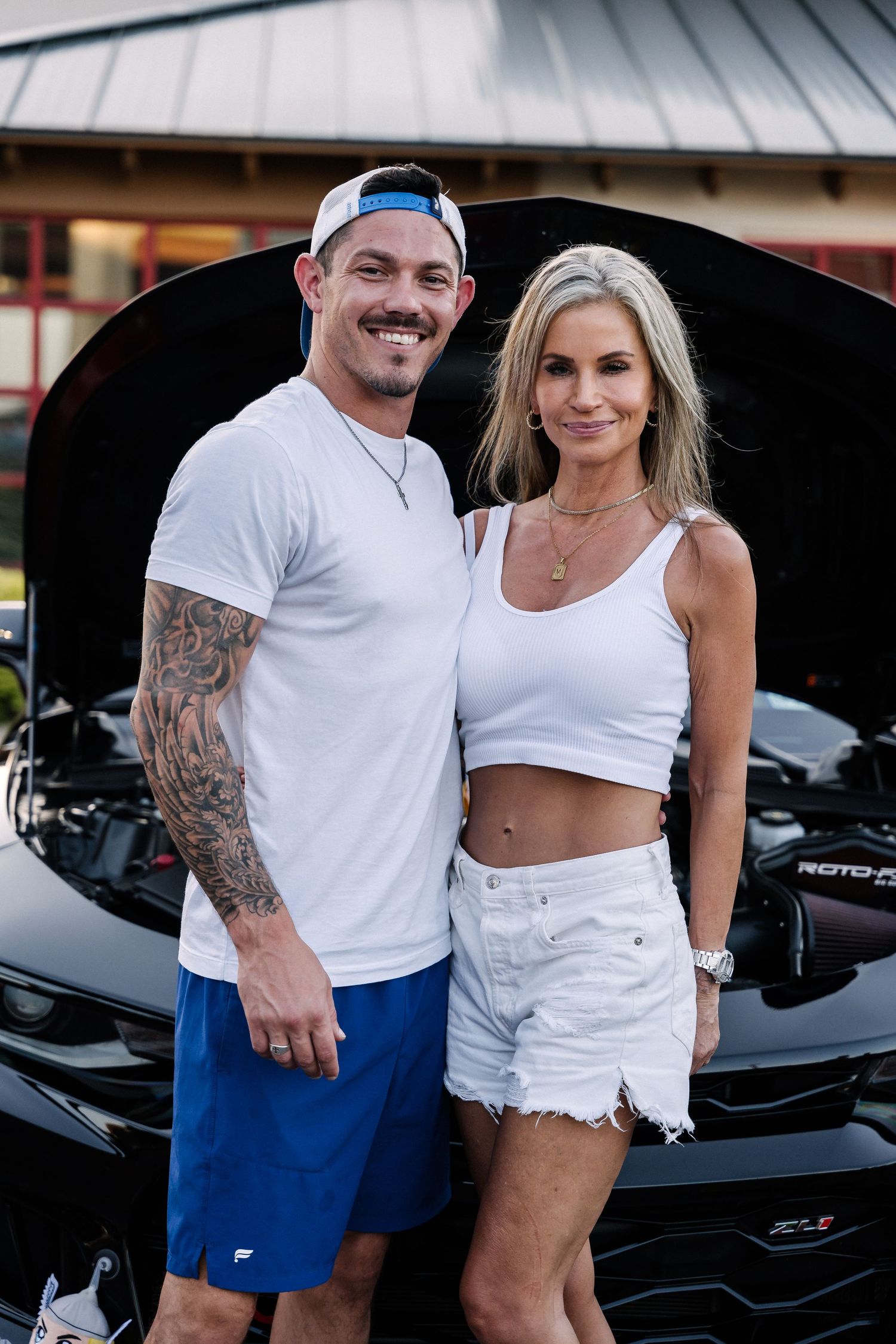A couple in white outfits posing together at a car meet with a black vehicle behind them.