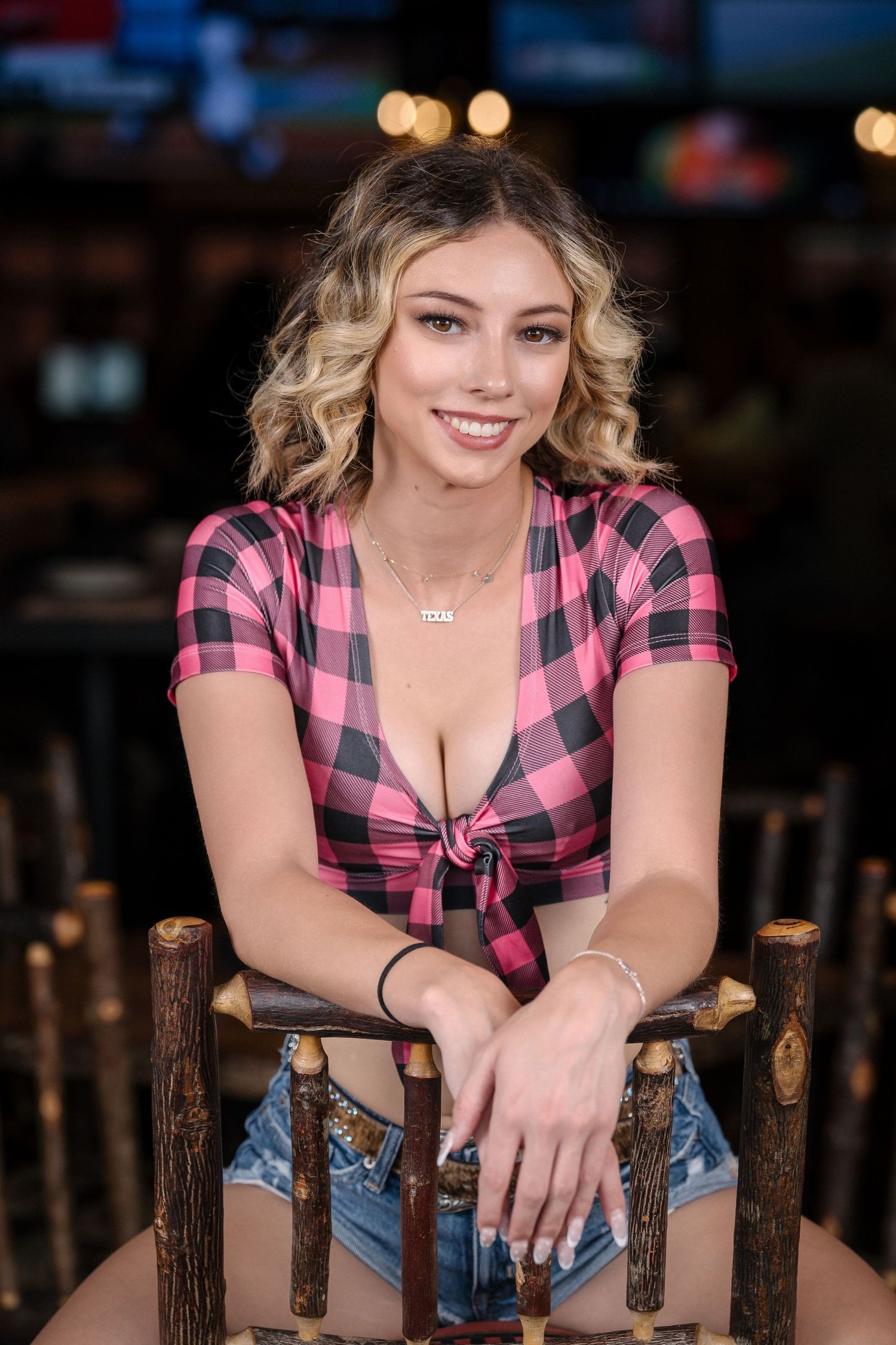 Casual portrait of model in pink plaid crop top sitting on rustic wooden chair.