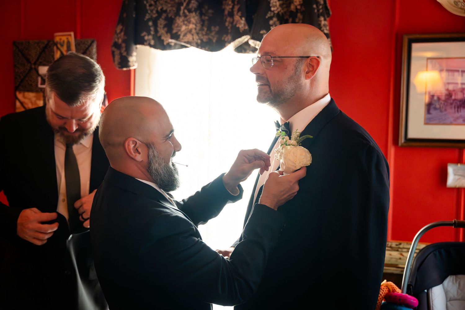 Groomsmen helping adjust boutonnieres and formal attire in dimly lit room with red walls.