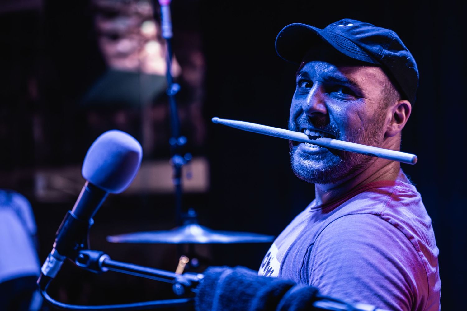 Drummer performs on stage with drumstick in mouth under blue stage lighting.