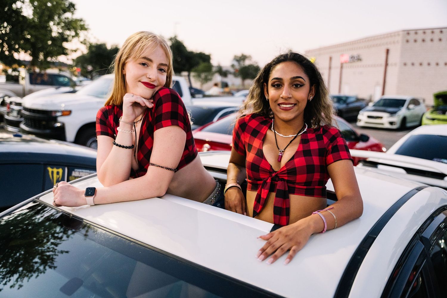 Two friends in matching red plaid tops smiling while sitting in white car at evening event.