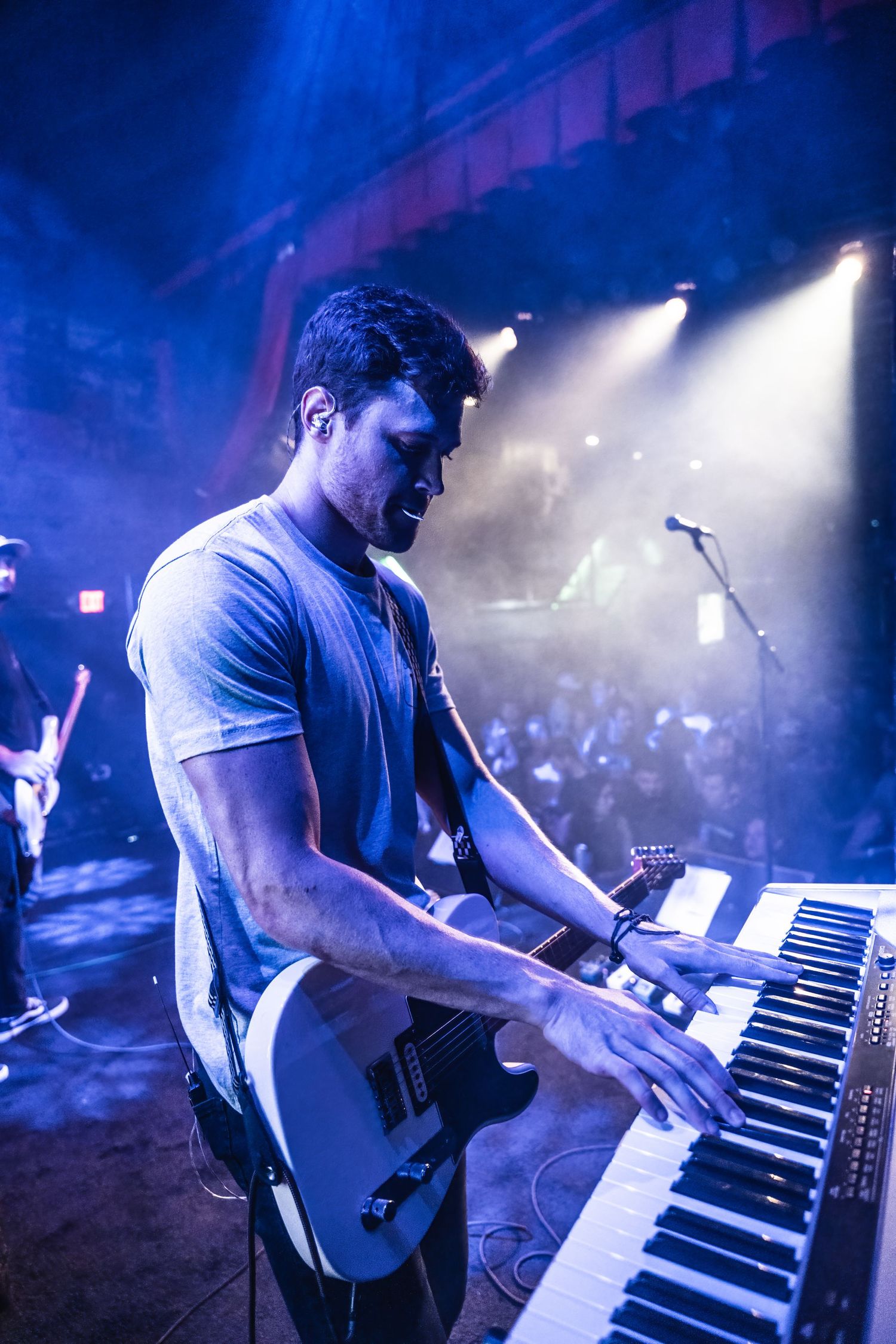 Musician playing keyboard and guitar simultaneously under dramatic blue stage lighting.