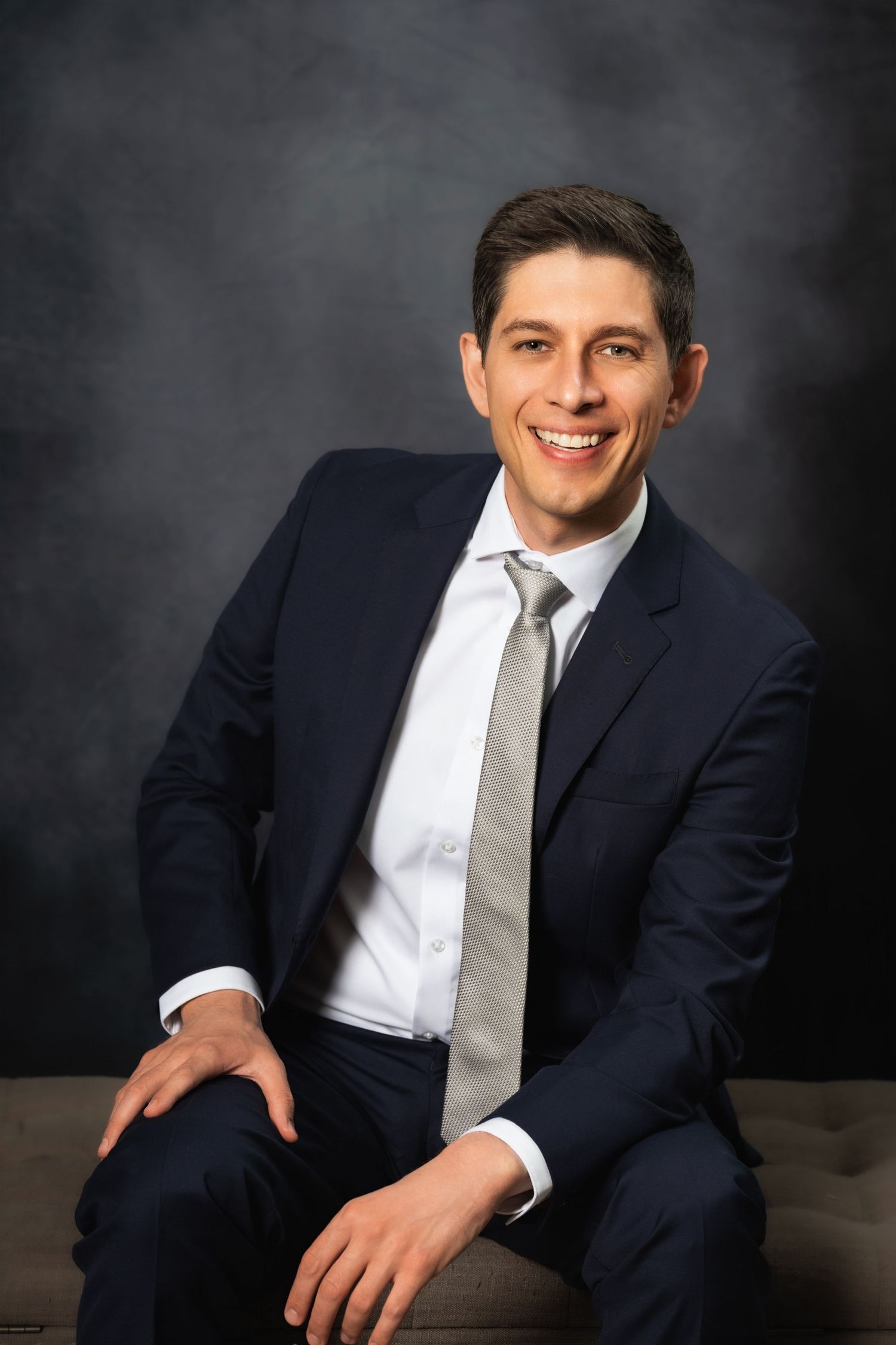 Casual business portrait of a seated professional in a navy suit with relaxed pose.