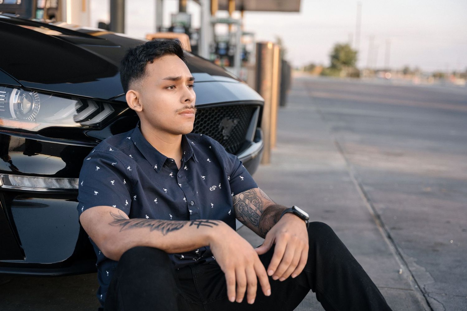 Person in dotted shirt sitting casually against black sports car at gas station.