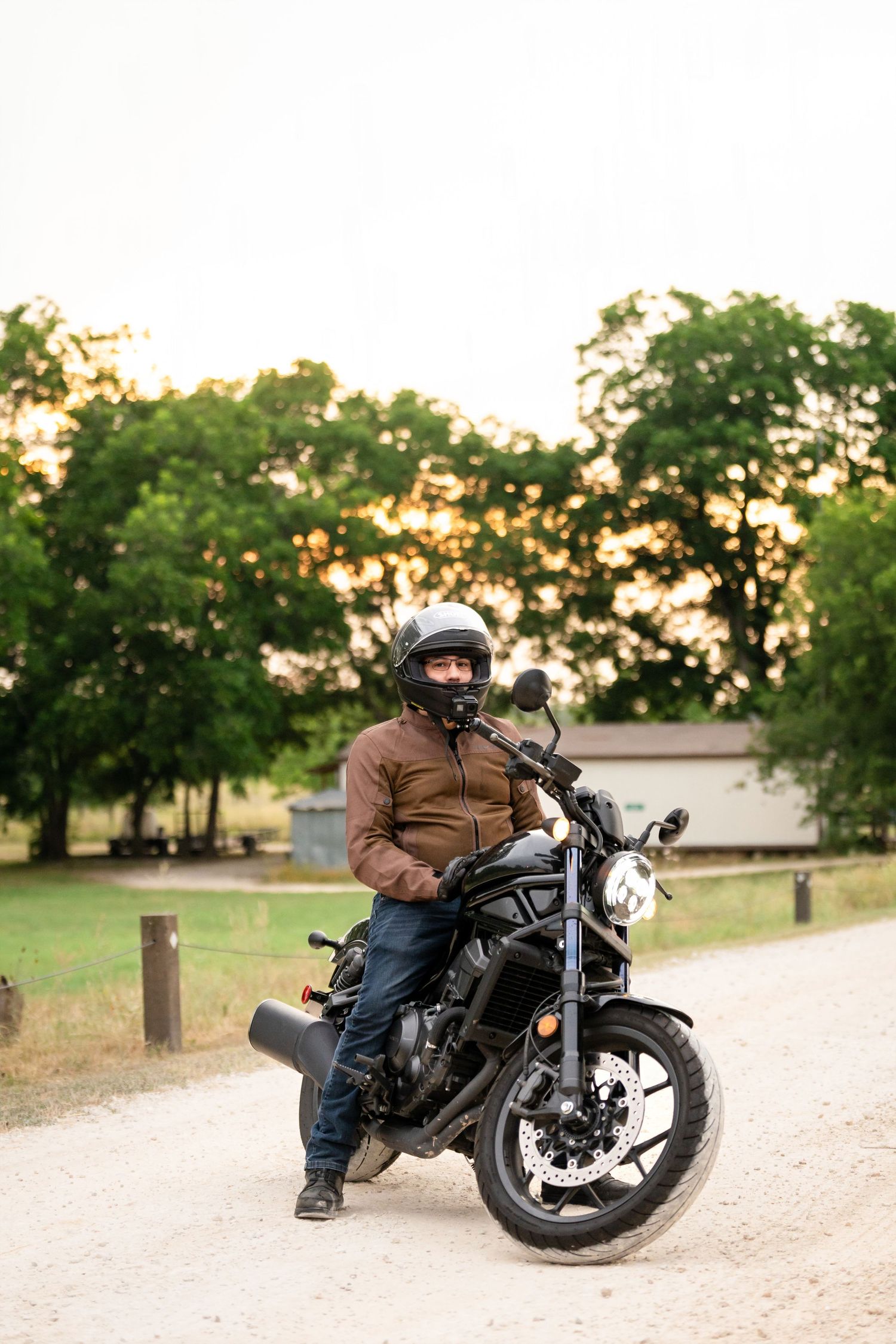 A motorcyclist in protective gear sits on a black motorcycle on a dirt path surrounded by trees.