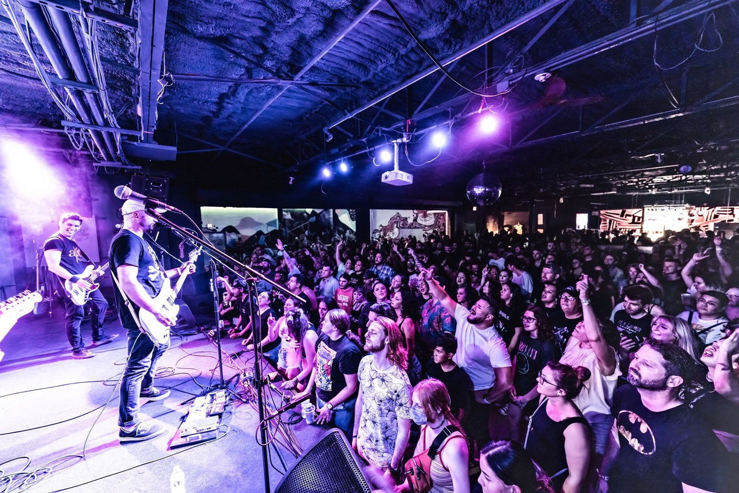 Wide shot of a packed music venue with performers on stage and enthusiastic crowd.