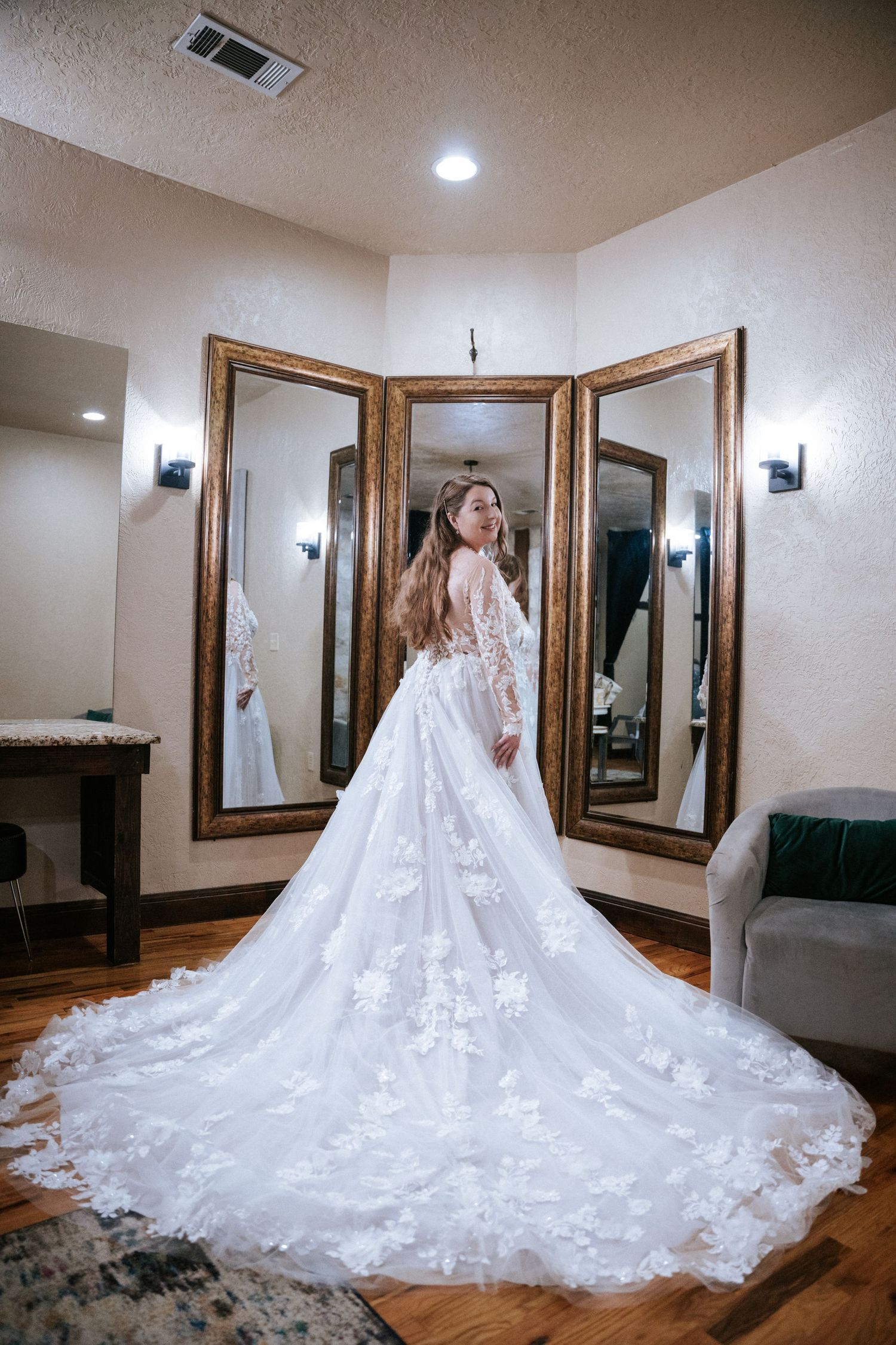 A wedding gown with intricate lace details and long train shown in a dressing room with three-panel mirror.