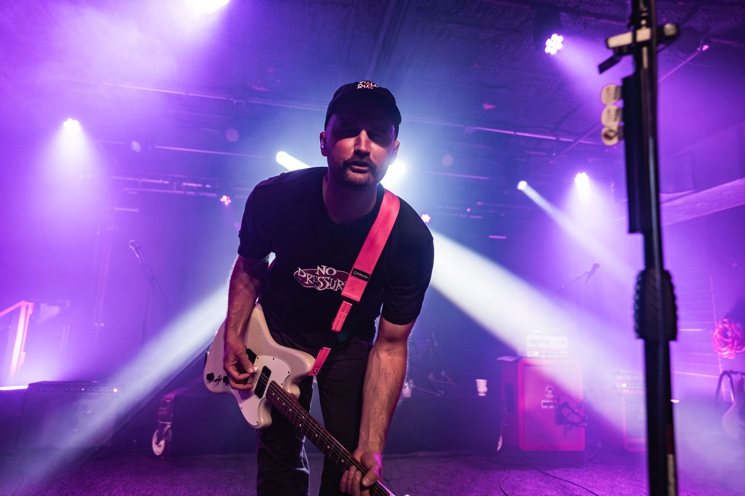 A performer plays bass guitar on stage beneath dramatic purple stage lighting during a concert performance.