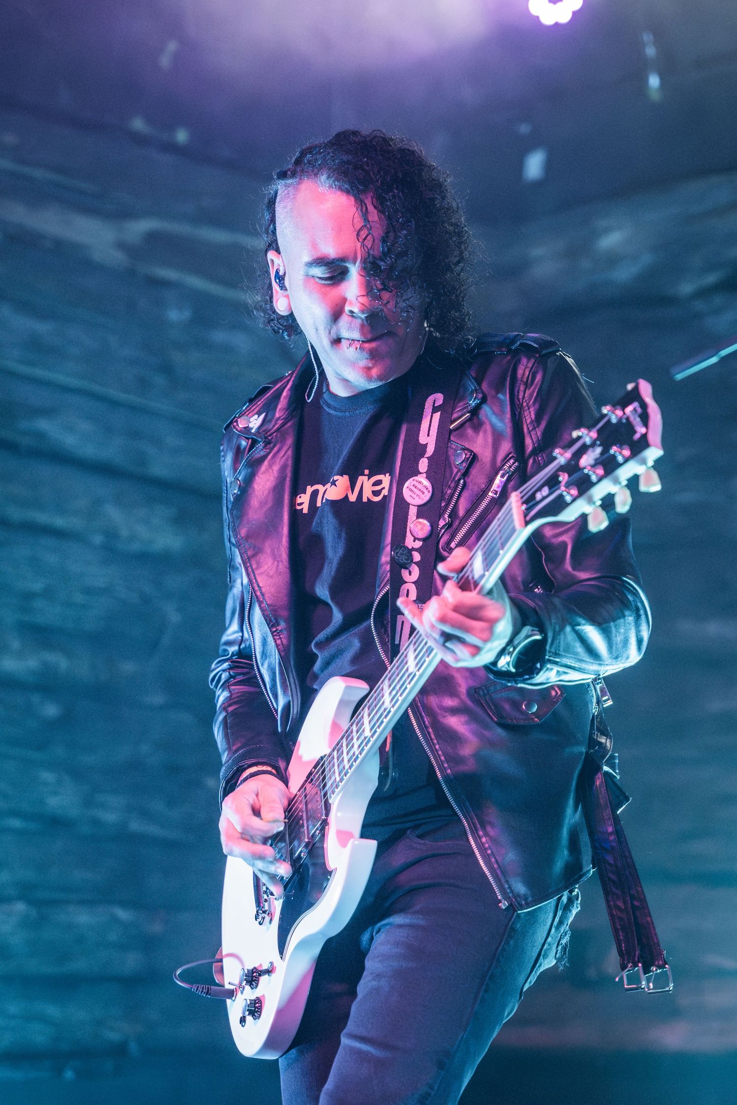 Guitarist in leather jacket performing under dramatic purple and blue stage lighting.