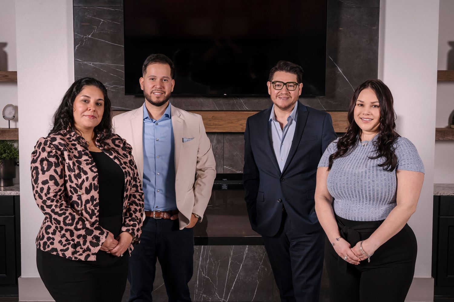 Four business professionals in professional attire stand together in front of a modern fireplace in an office setting.