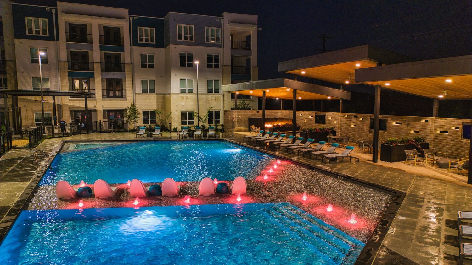 Swimming pool with glowing pink floating loungers and modern apartment buildings in the background.