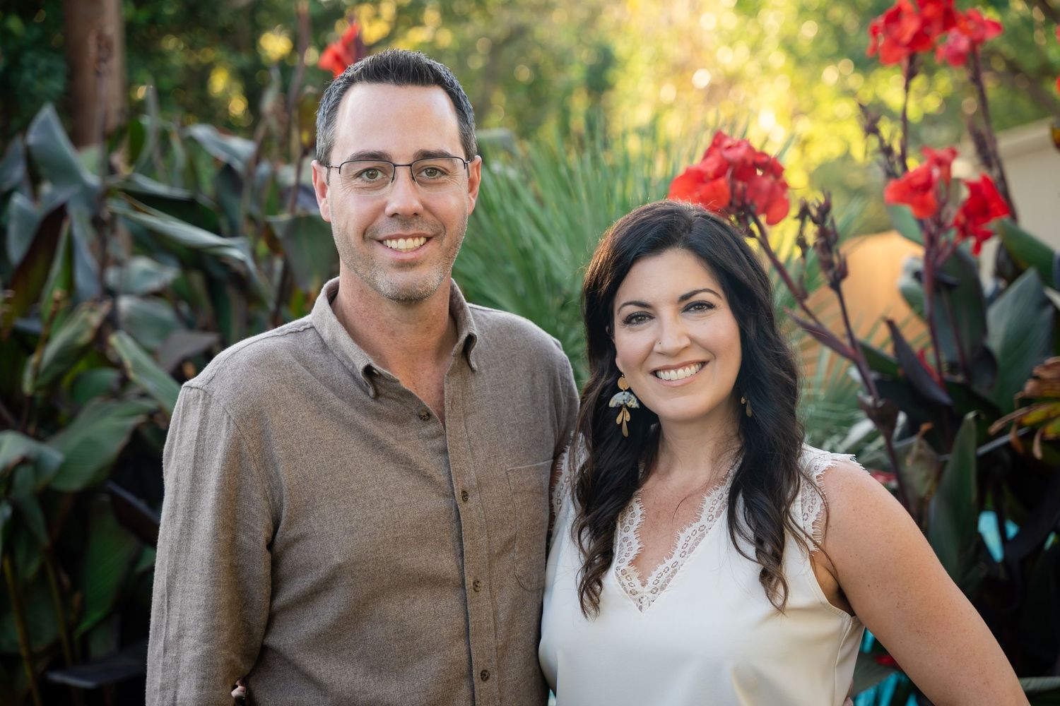 Couple poses in garden setting with red flowers and lush greenery.