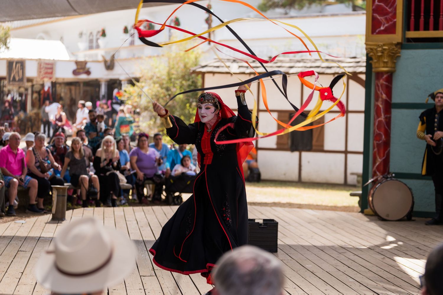 A performer in black costume twirls red ribbons on stage during a Renaissance festival performance.