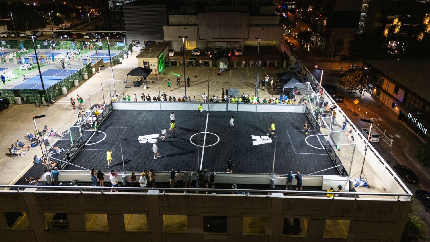 Birds-eye view of two illuminated rooftop soccer fields during nighttime matches.