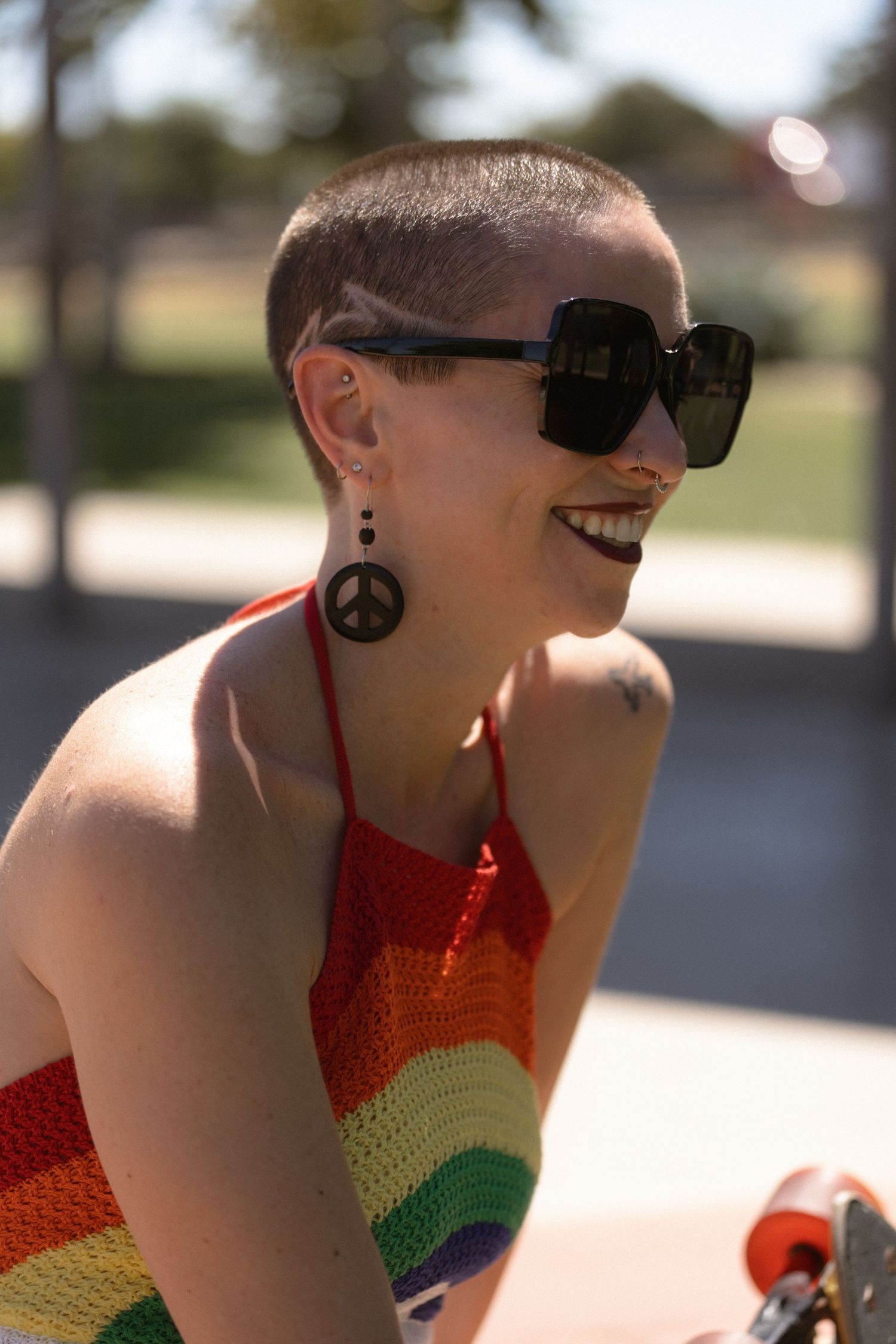 A person in sunglasses and peace sign earrings wearing a rainbow colored halter top smiles outdoors.