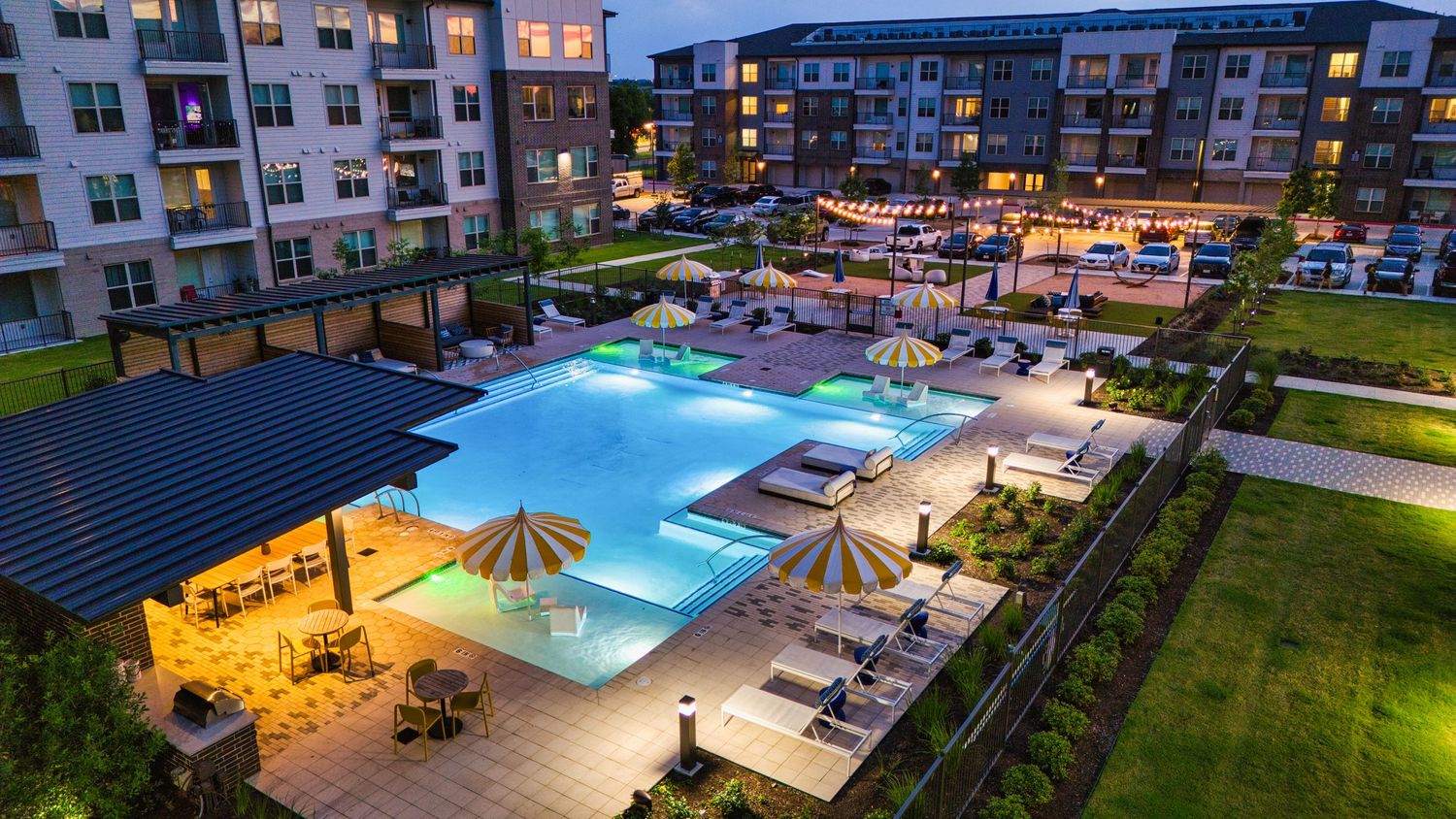 Luxury apartment complex pool area illuminated at dusk with lounge chairs and umbrellas surrounding the water.