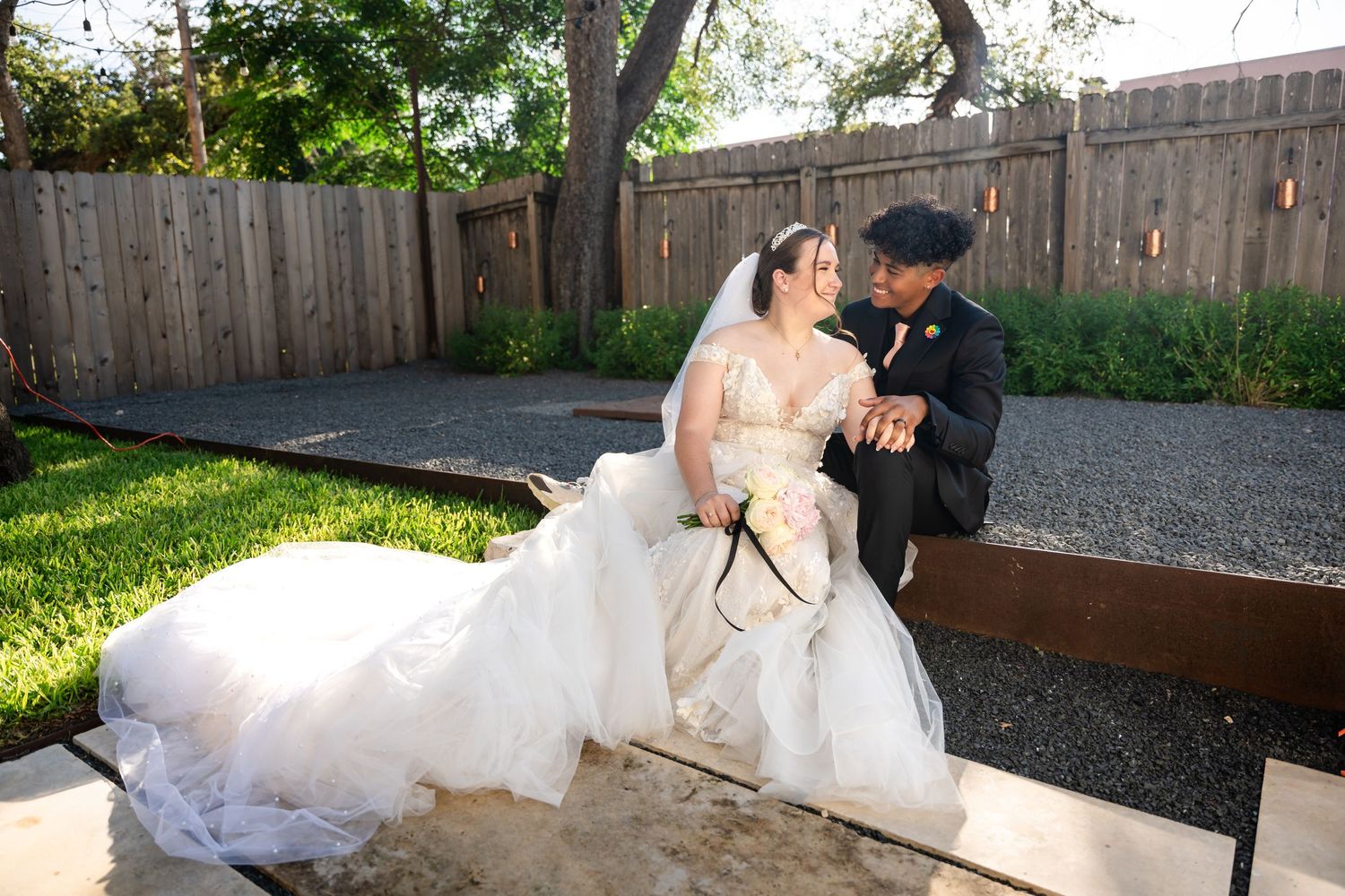 Bride in white gown and groom in dark suit sitting together outdoors by wooden fence.