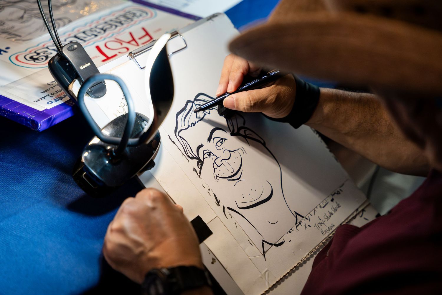Artist sketching a portrait with a pen on paper while wearing headphones at a drawing table with a blue cloth surface.