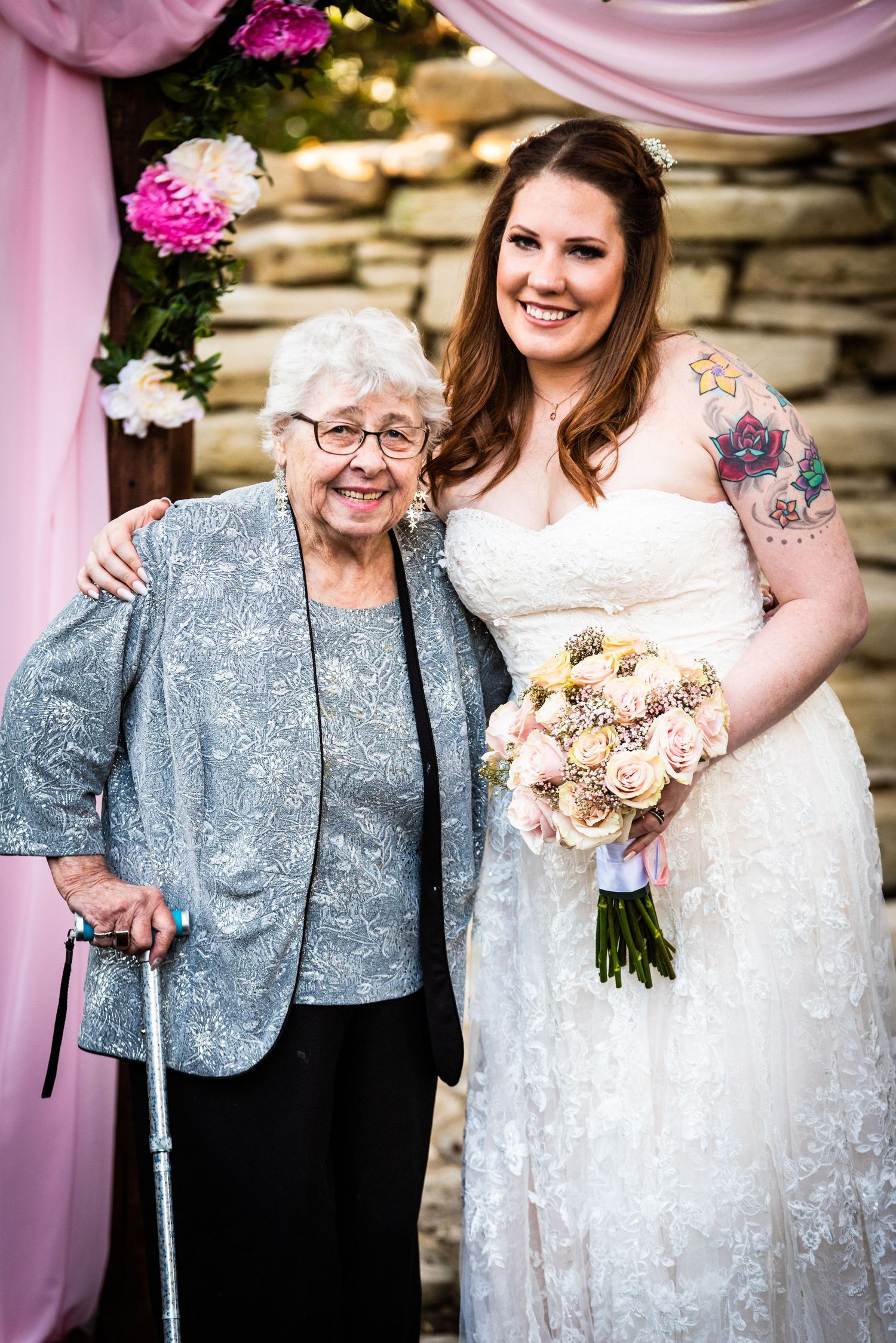 Bride in lace dress poses with elderly relative in sparkly jacket at wedding ceremony.
