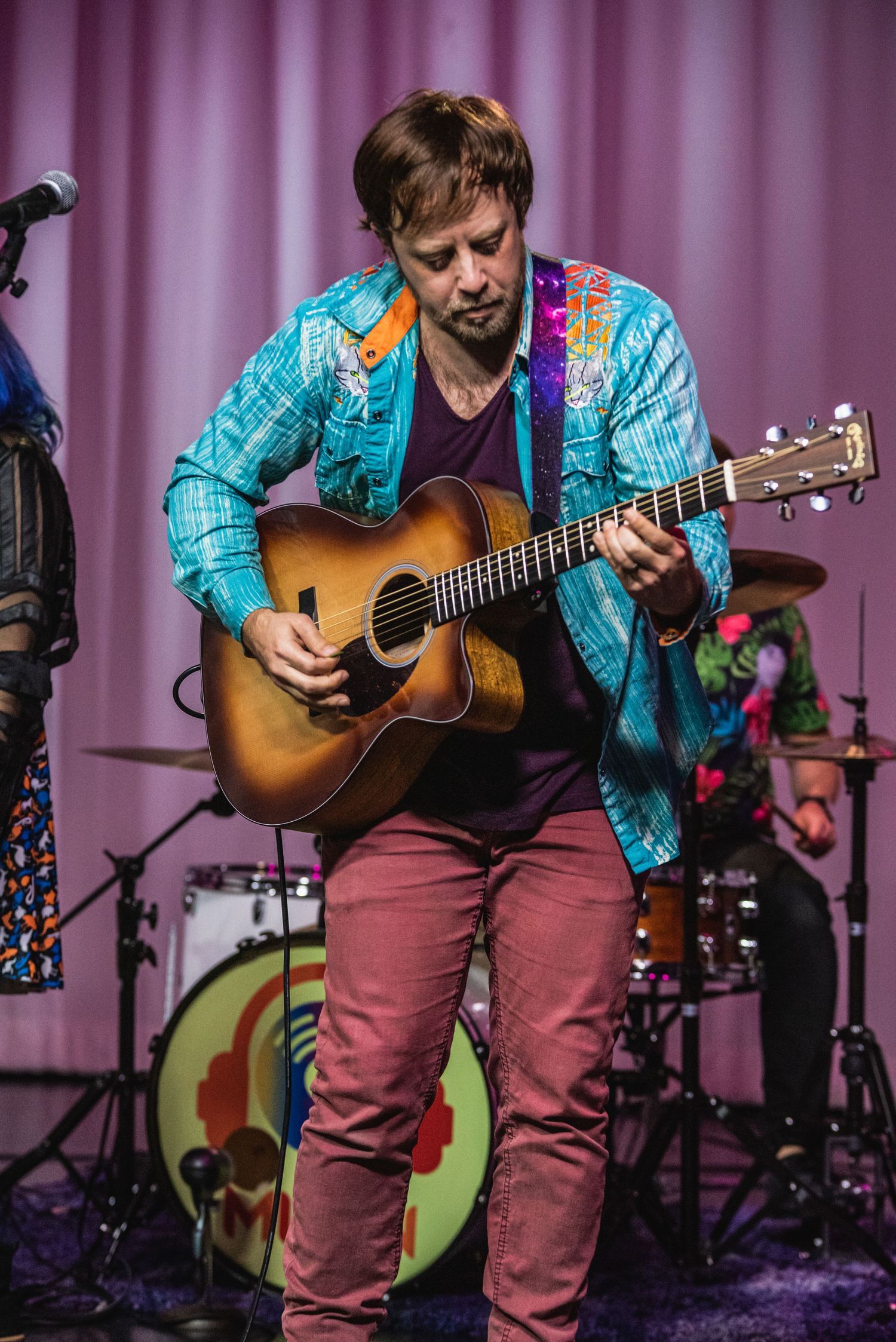 Guitarist in turquoise jacket and red pants plays acoustic guitar during stage performance.