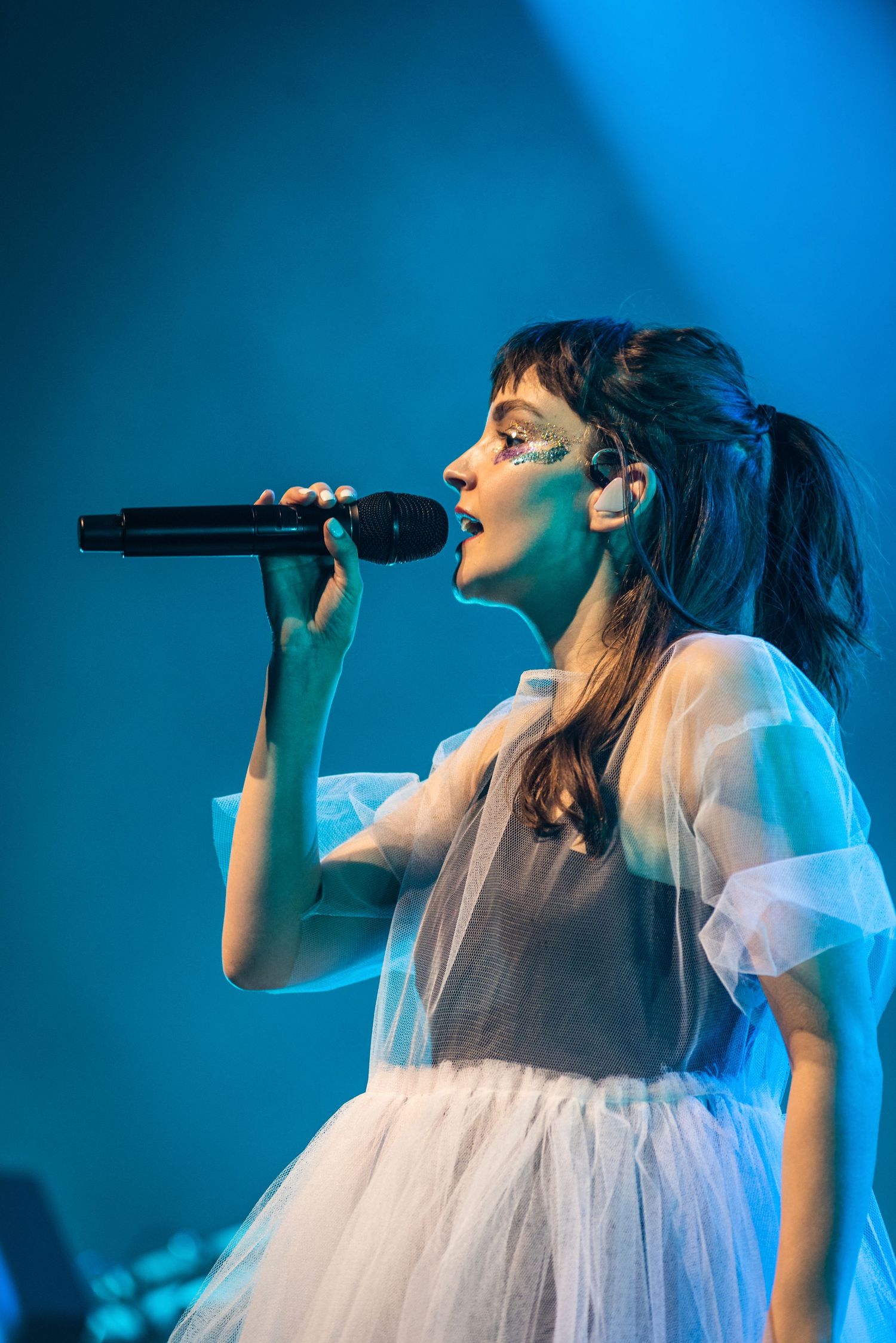 Concert performer in a white tulle dress sings into a microphone against a blue-lit background.