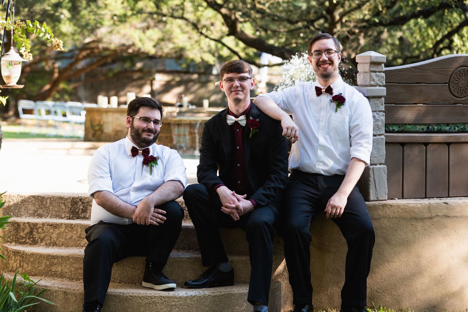 Three groomsmen in formal attire sit casually on stone steps at an outdoor wedding venue.