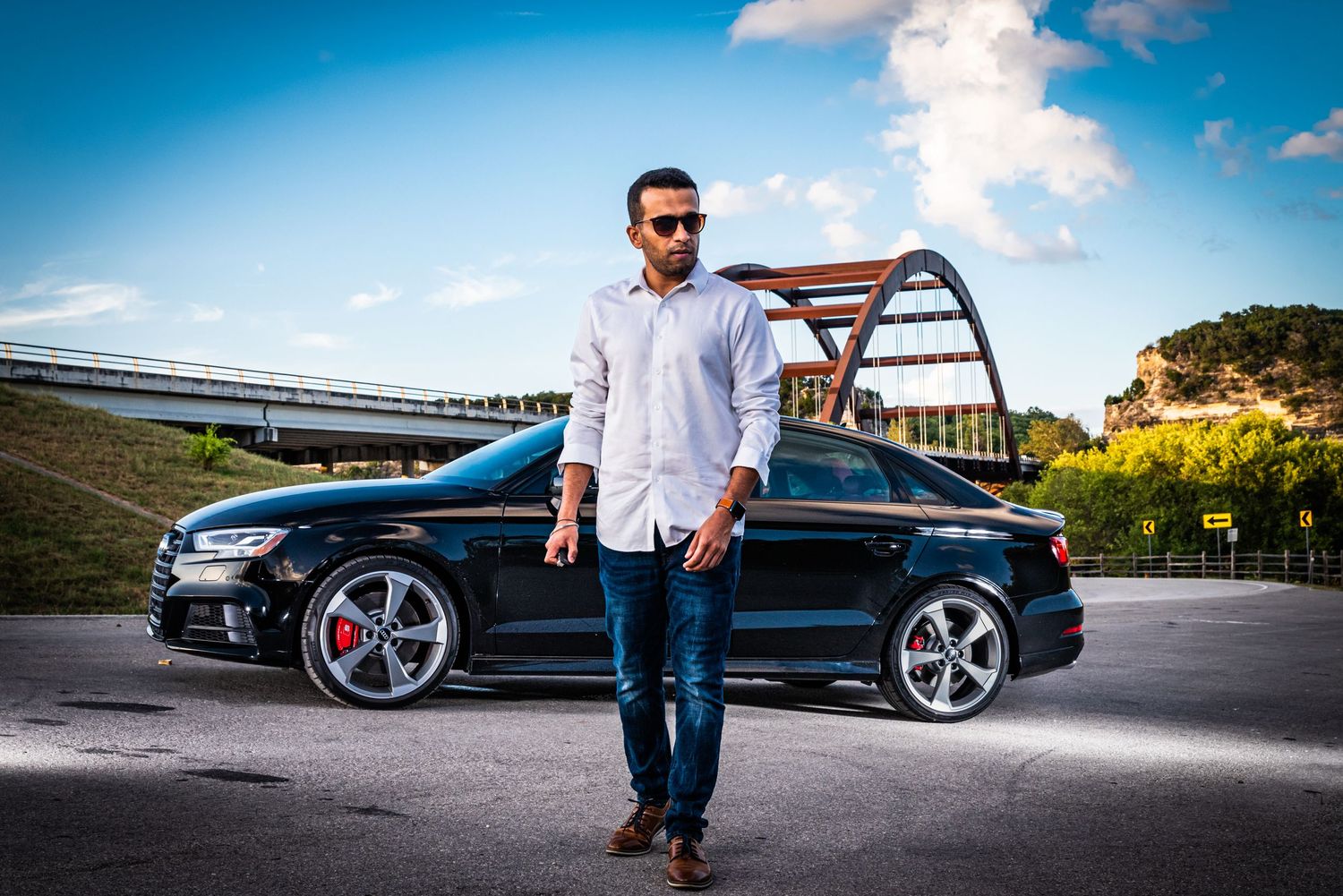 A sleek black Audi S3 parked in front of a steel arch bridge during golden hour with warm lighting.