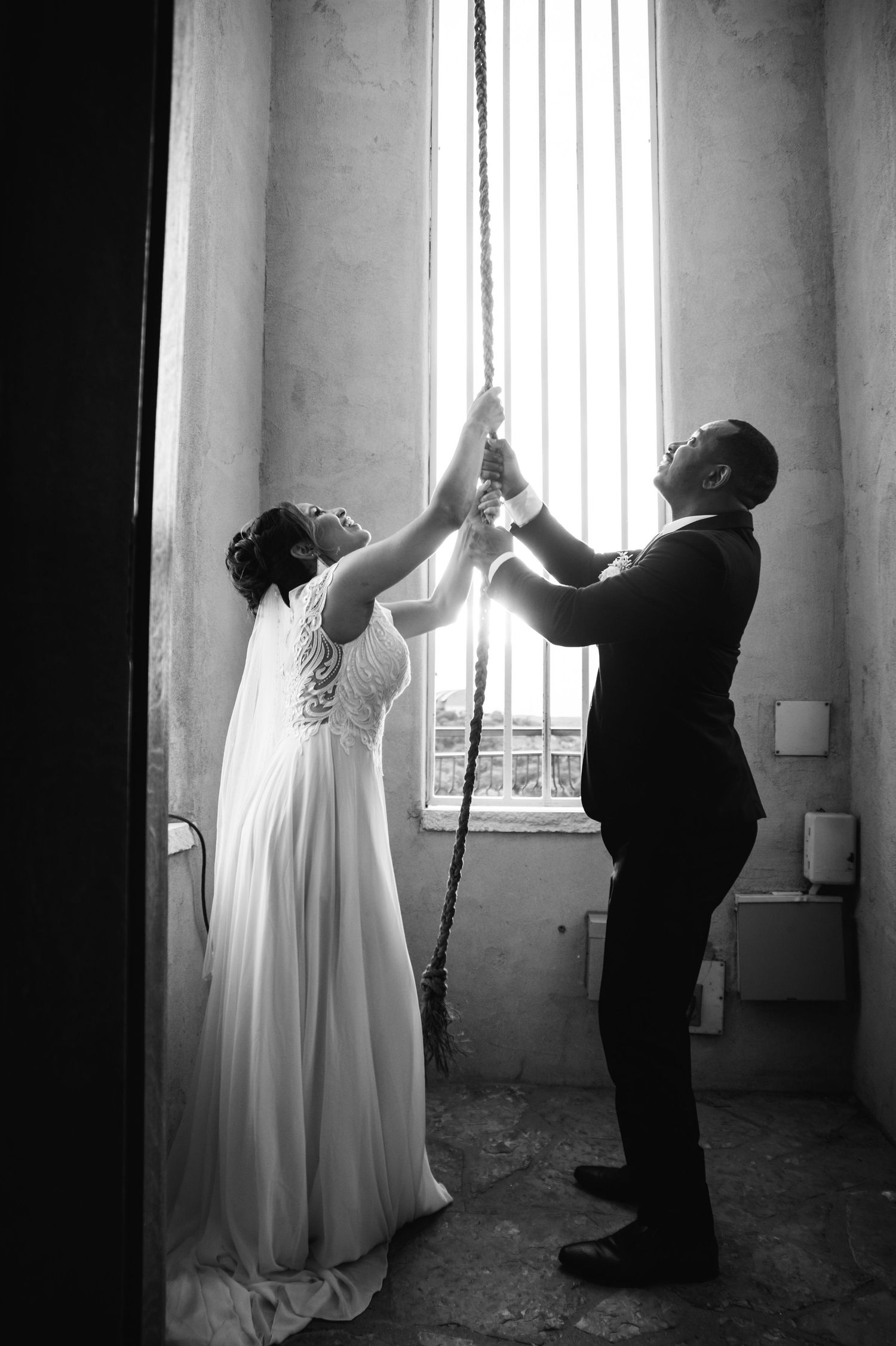 A dramatic black and white wedding portrait showing a couple reaching up toward streaming window light.