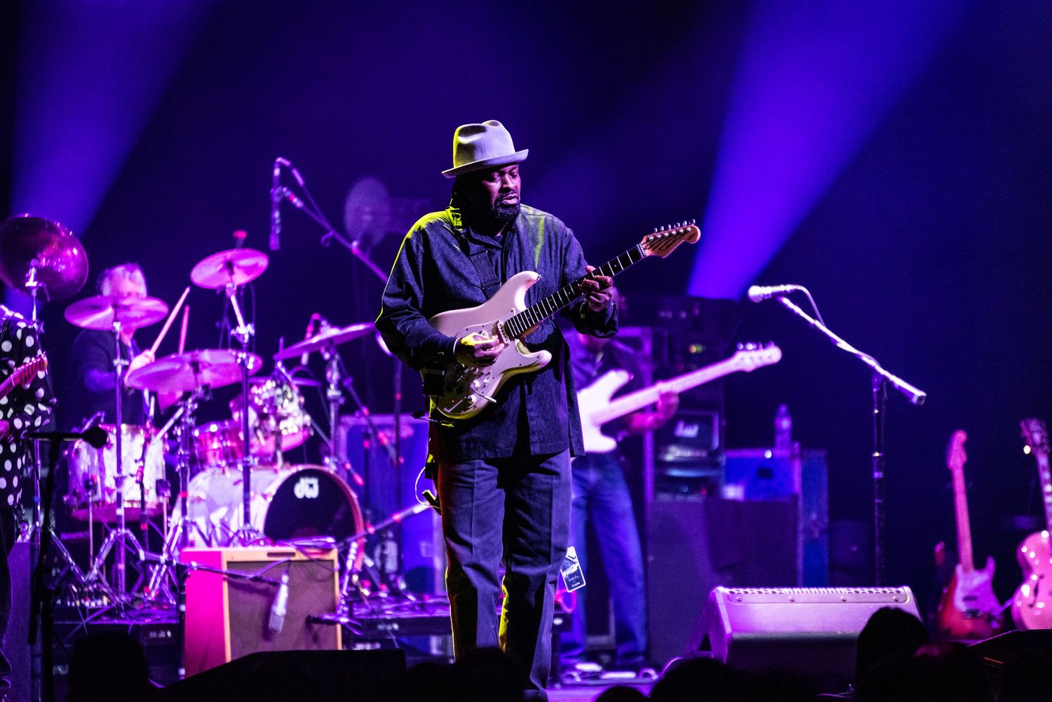 Concert stage lit with purple lights showing performer with guitar and drum kit in background.