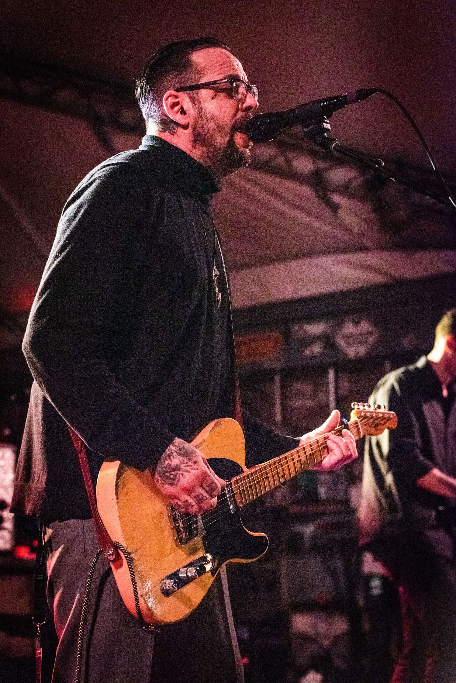Musician playing a Telecaster guitar and singing into microphone during energetic stage performance.