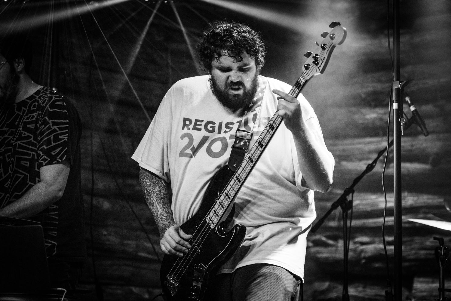 Black and white shot of bass player wearing Register To Vote shirt performing on stage.