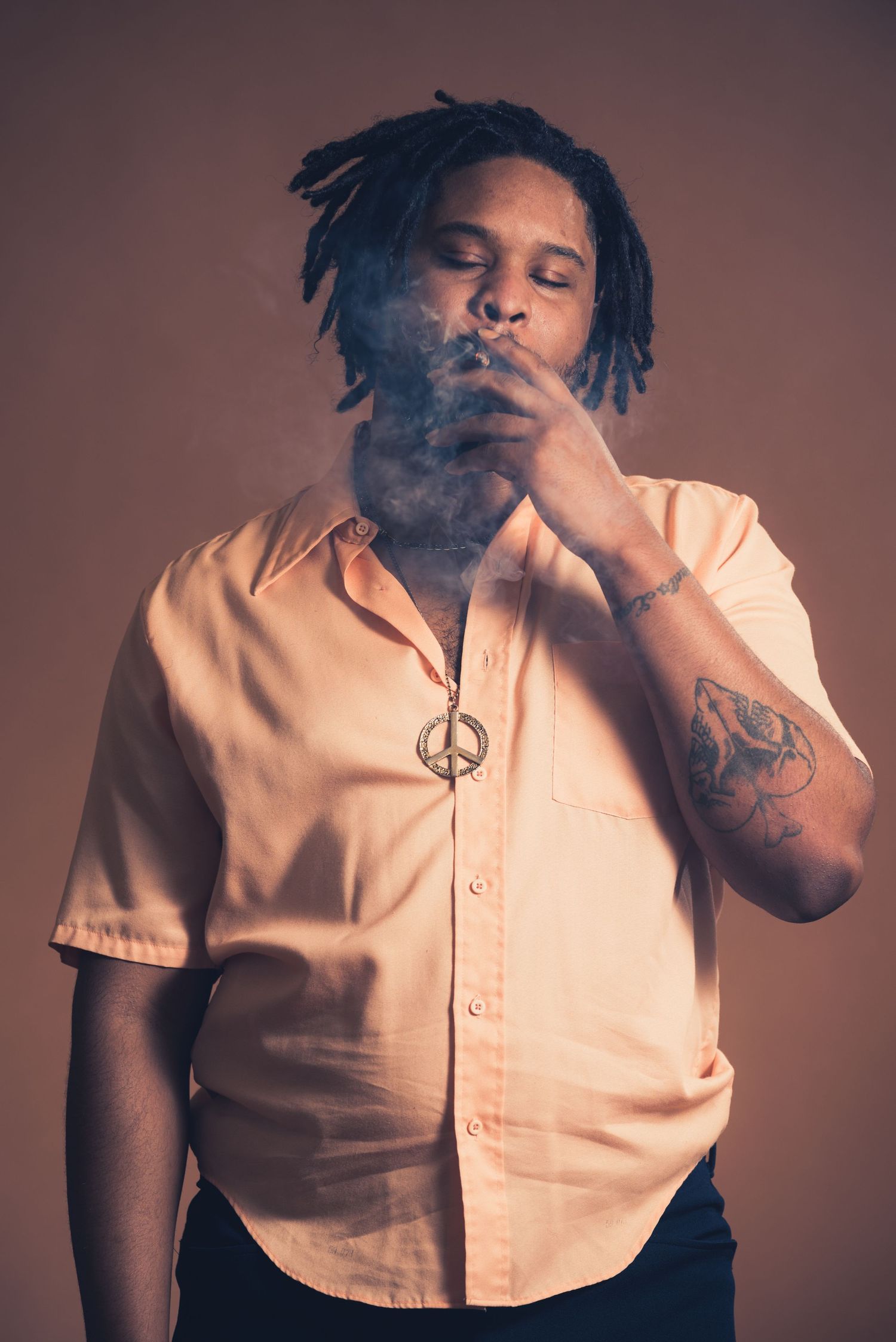 Person in peach shirt with dreadlocks exhaling smoke while wearing a peace sign necklace.