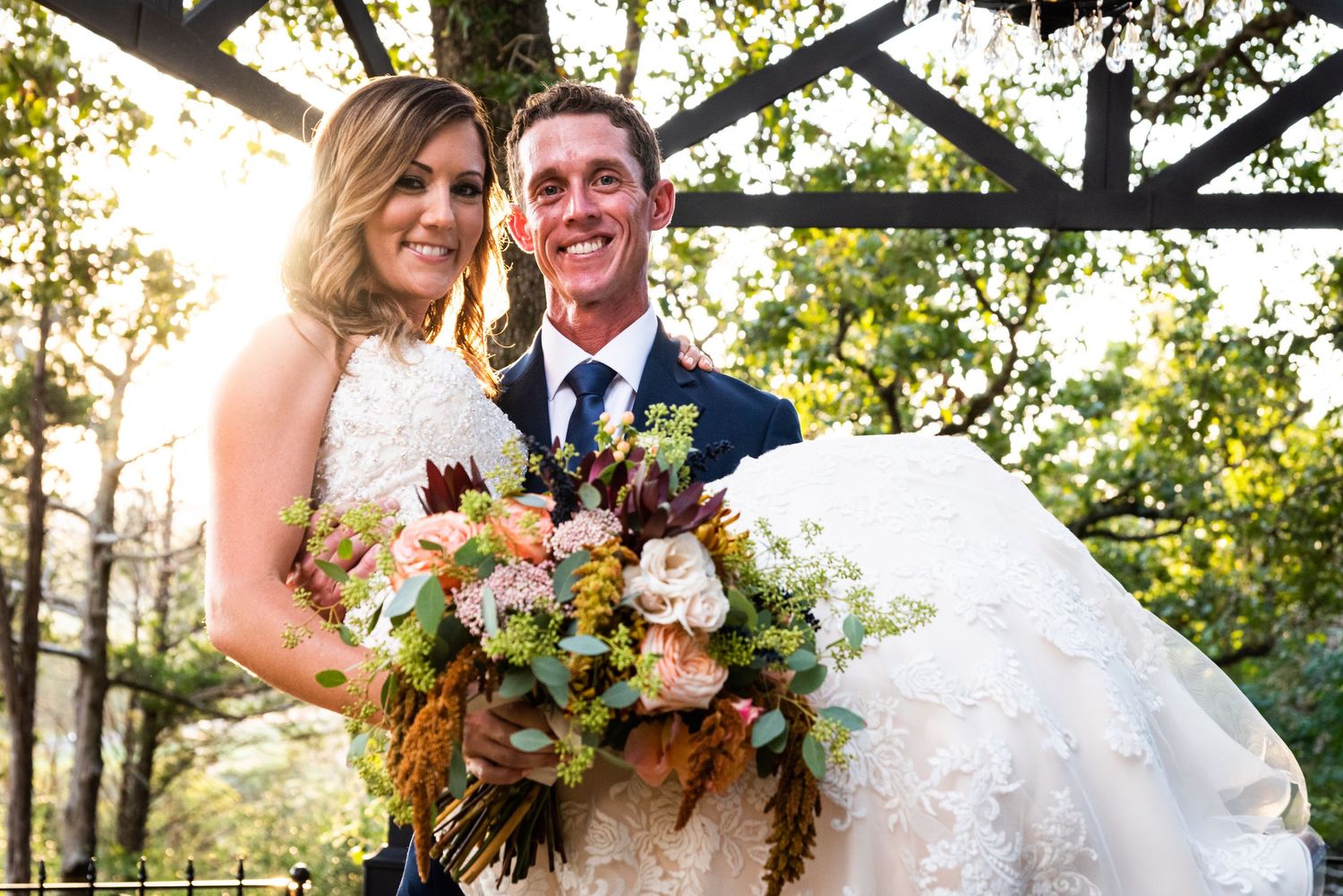 A newlywed couple poses with a lush fall-colored bouquet under an outdoor pergola.