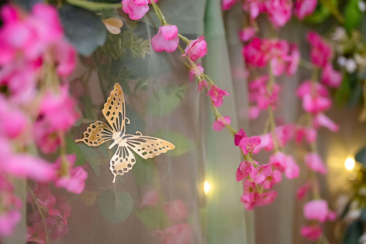 Golden butterfly ornament hanging among pink flowers and green fabric with soft bokeh lights in background.