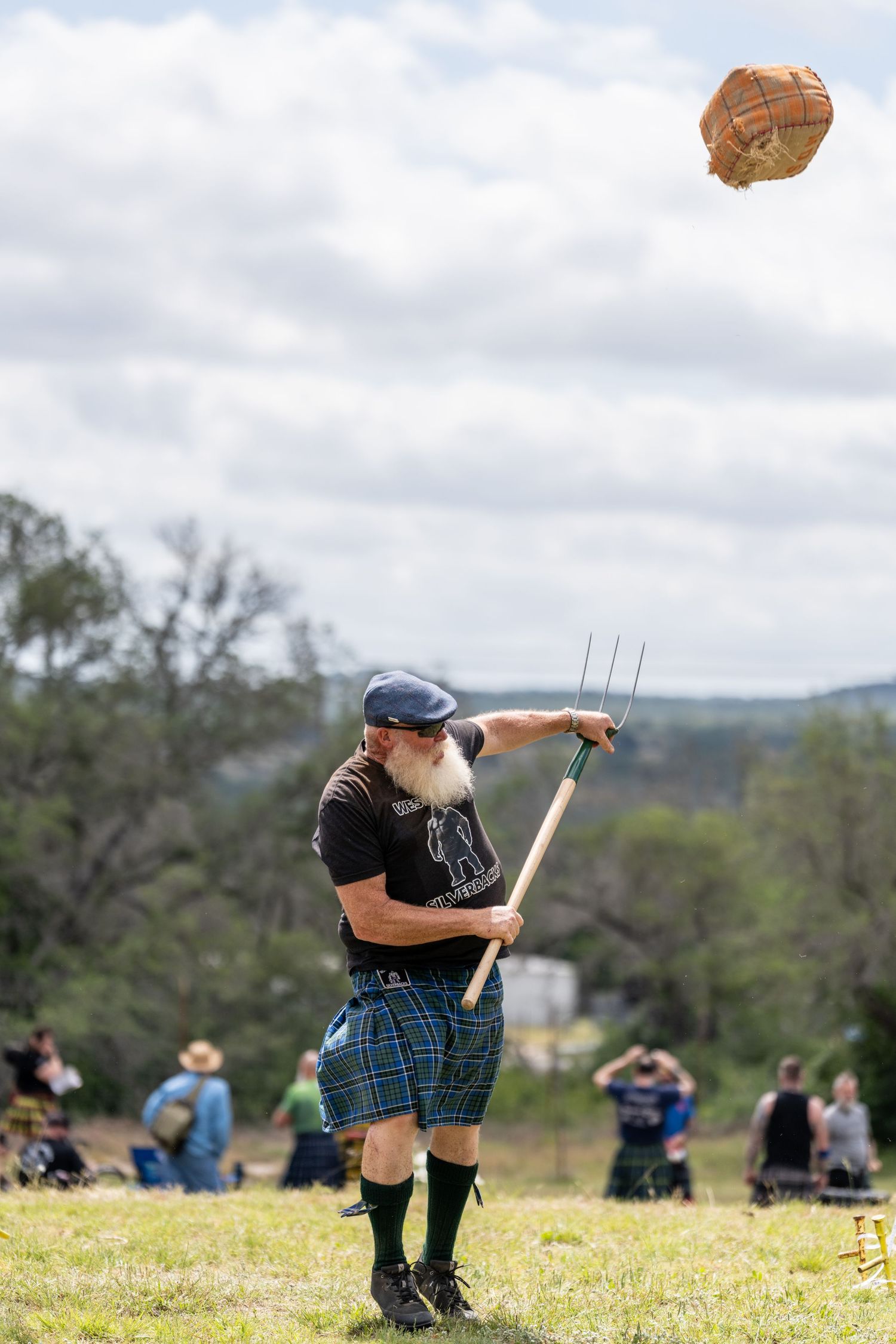 Bearded man in tartan kilt throws sheaf with pitchfork at Scottish Highland Games outdoor competition.
