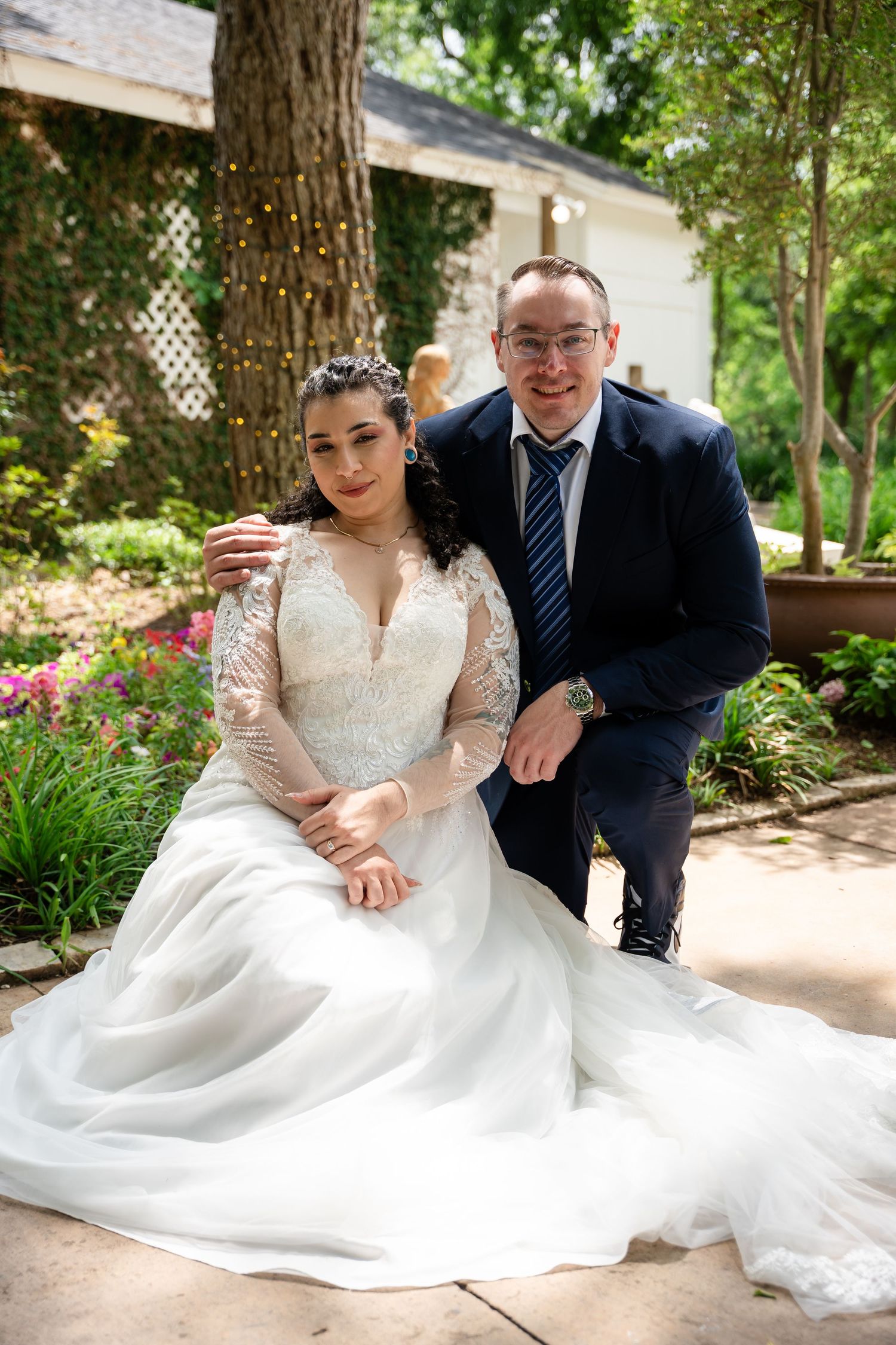 Bride in lace gown and groom in navy suit pose together in a lush garden wedding setting.
