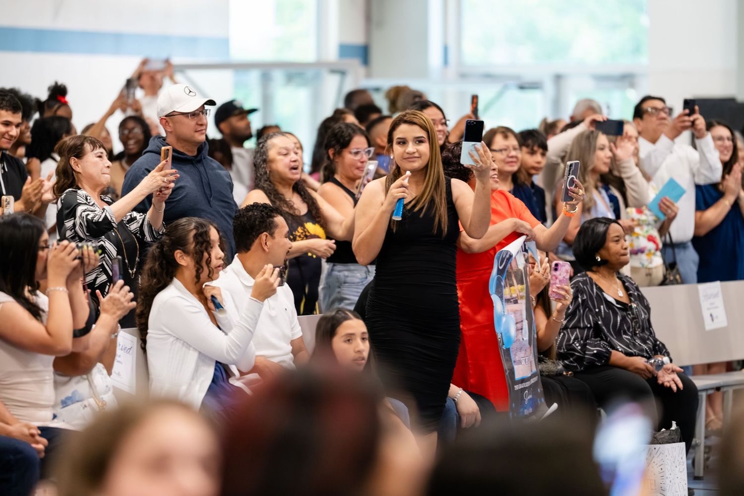 Crowd of diverse people at an indoor event, many holding phones to capture photos, with a woman in blue dress standing out.