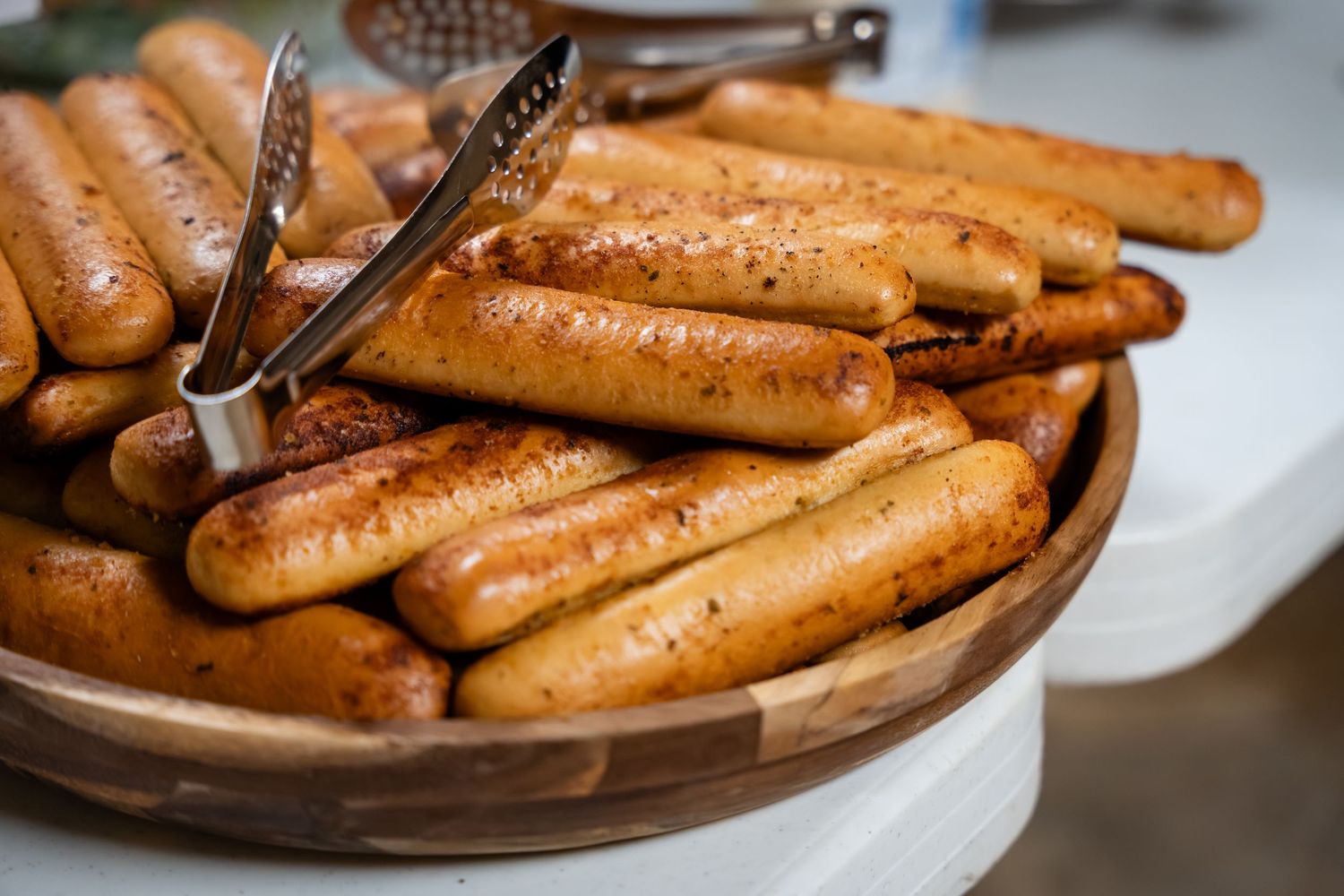 Grilled sausages piled in a wooden bowl with metal tongs, golden-brown and seasoned, perfect for a buffet or BBQ event.