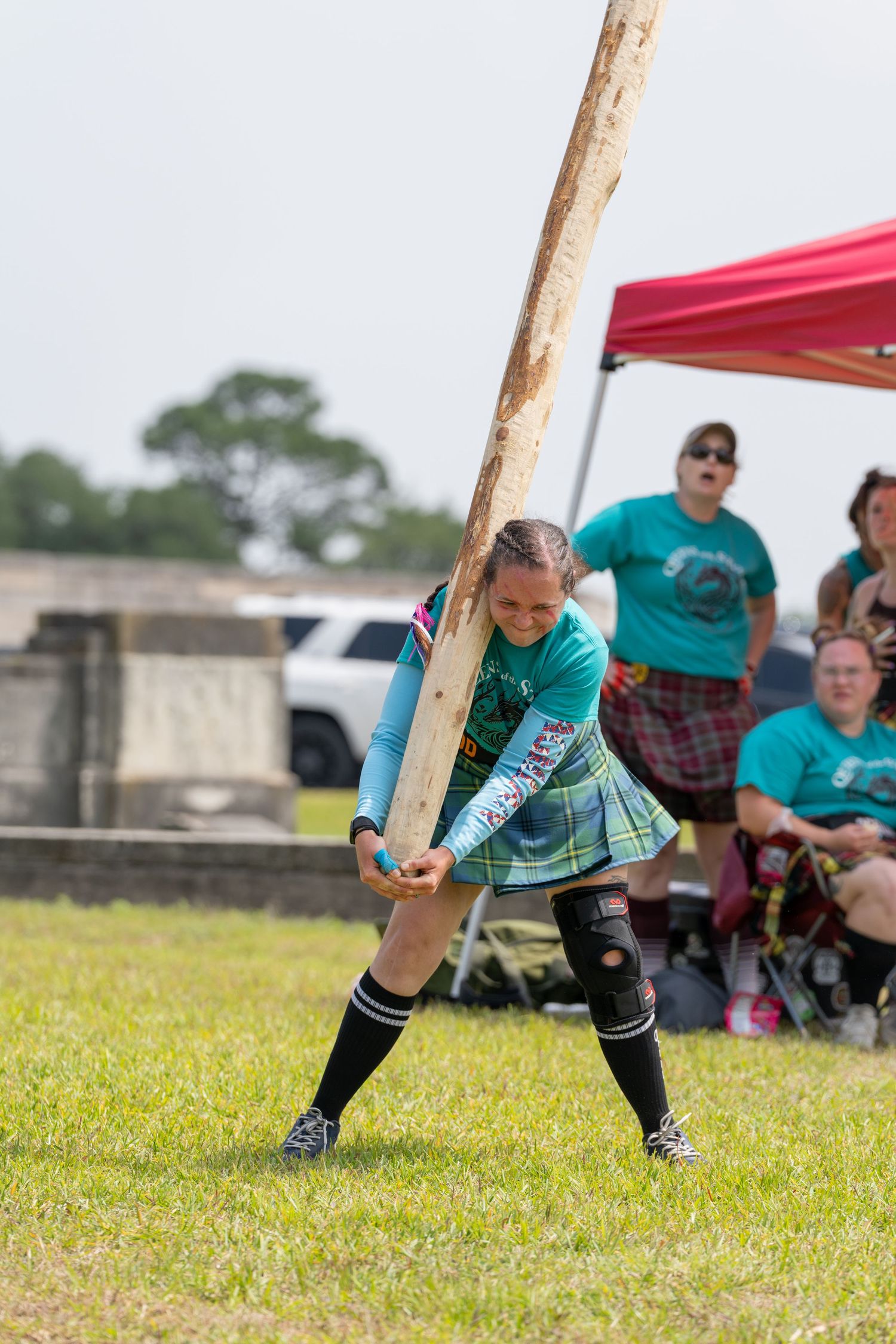 Woman competing in caber toss at Highland Games, lifting large wooden pole on green grass field.