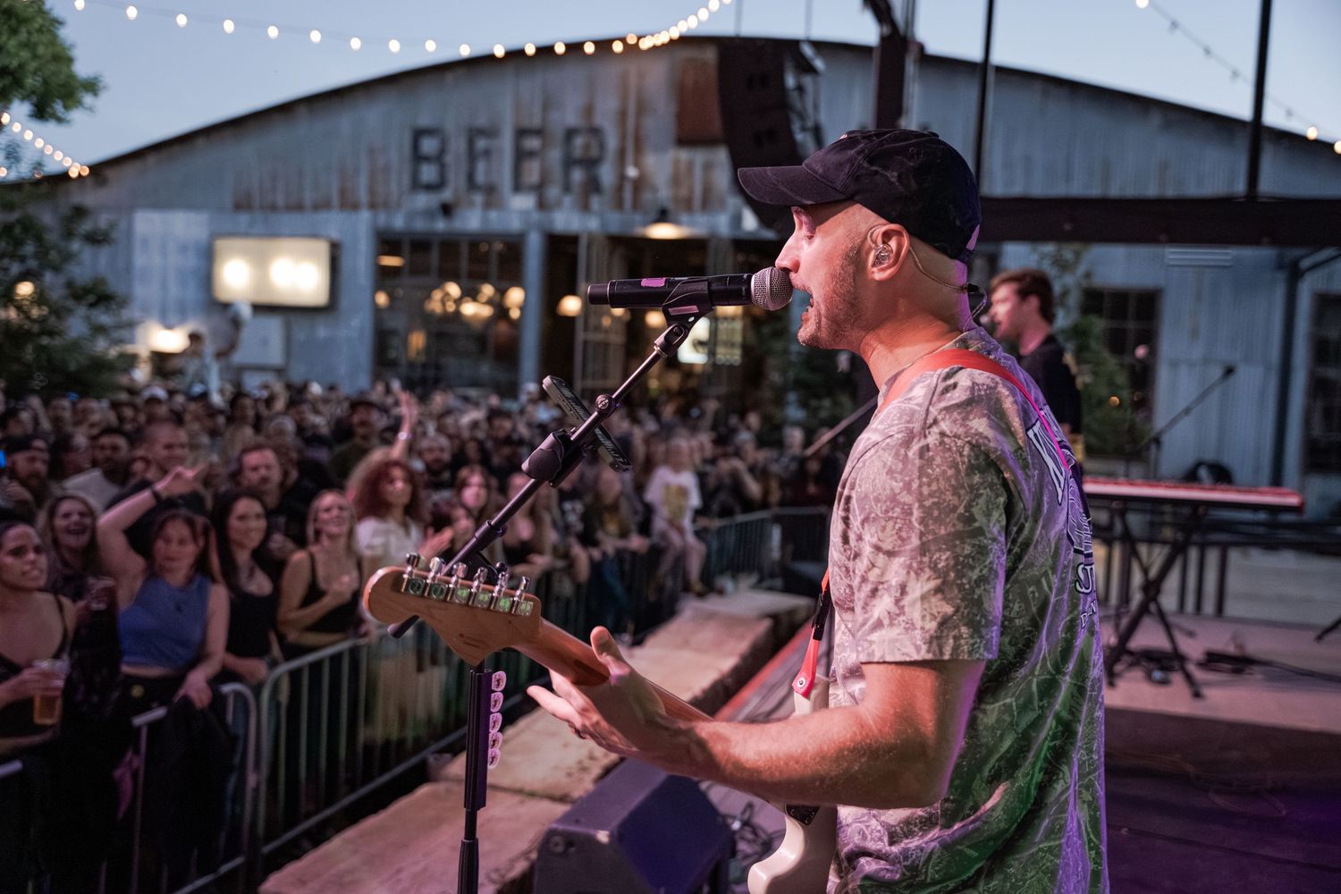 Guitarist performing on outdoor stage at a beer venue concert with a large crowd of fans gathered at dusk.